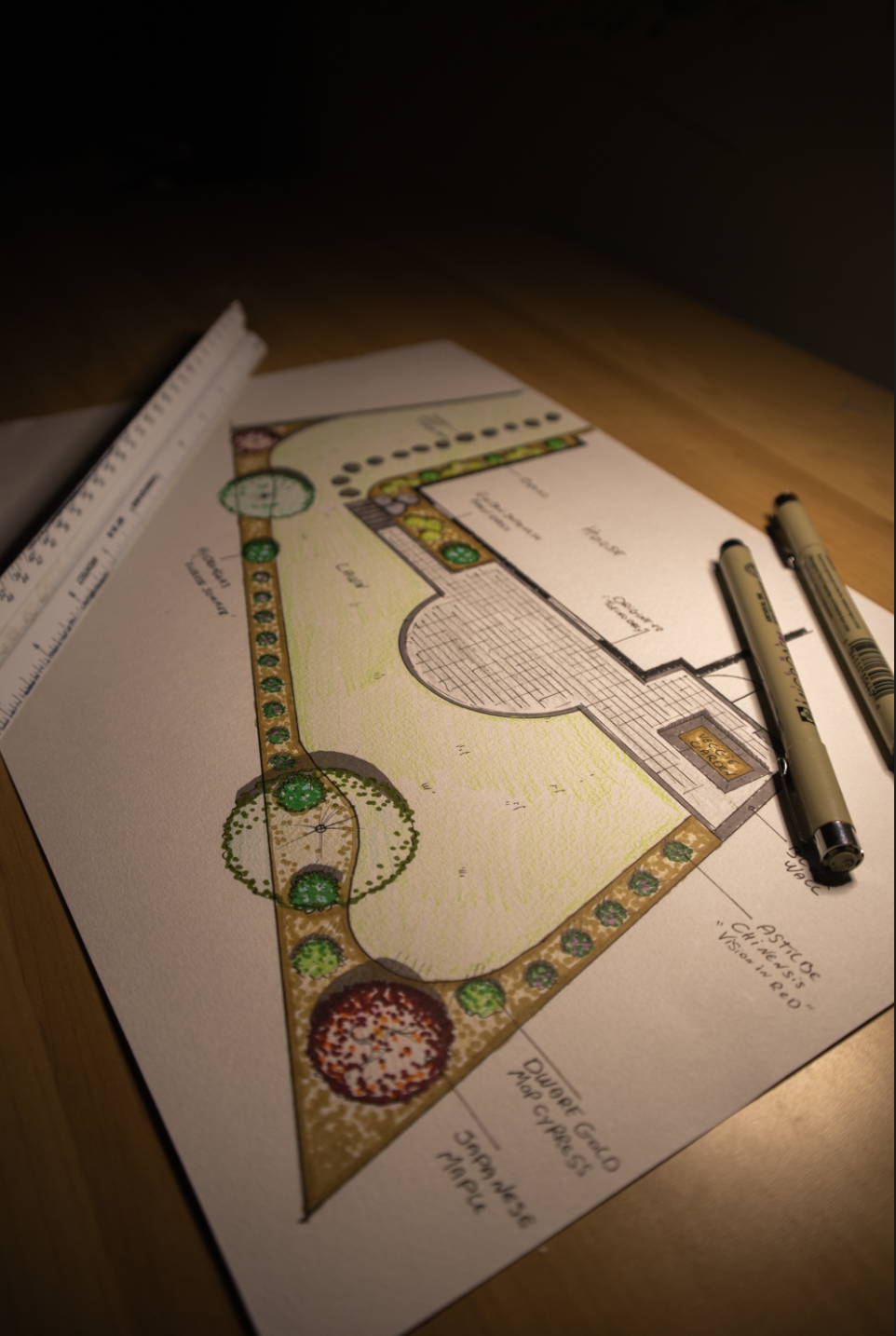 Landscape design plan on paper with trees, pathways, and a building layout, placed on a wooden table with pens and a ruler.