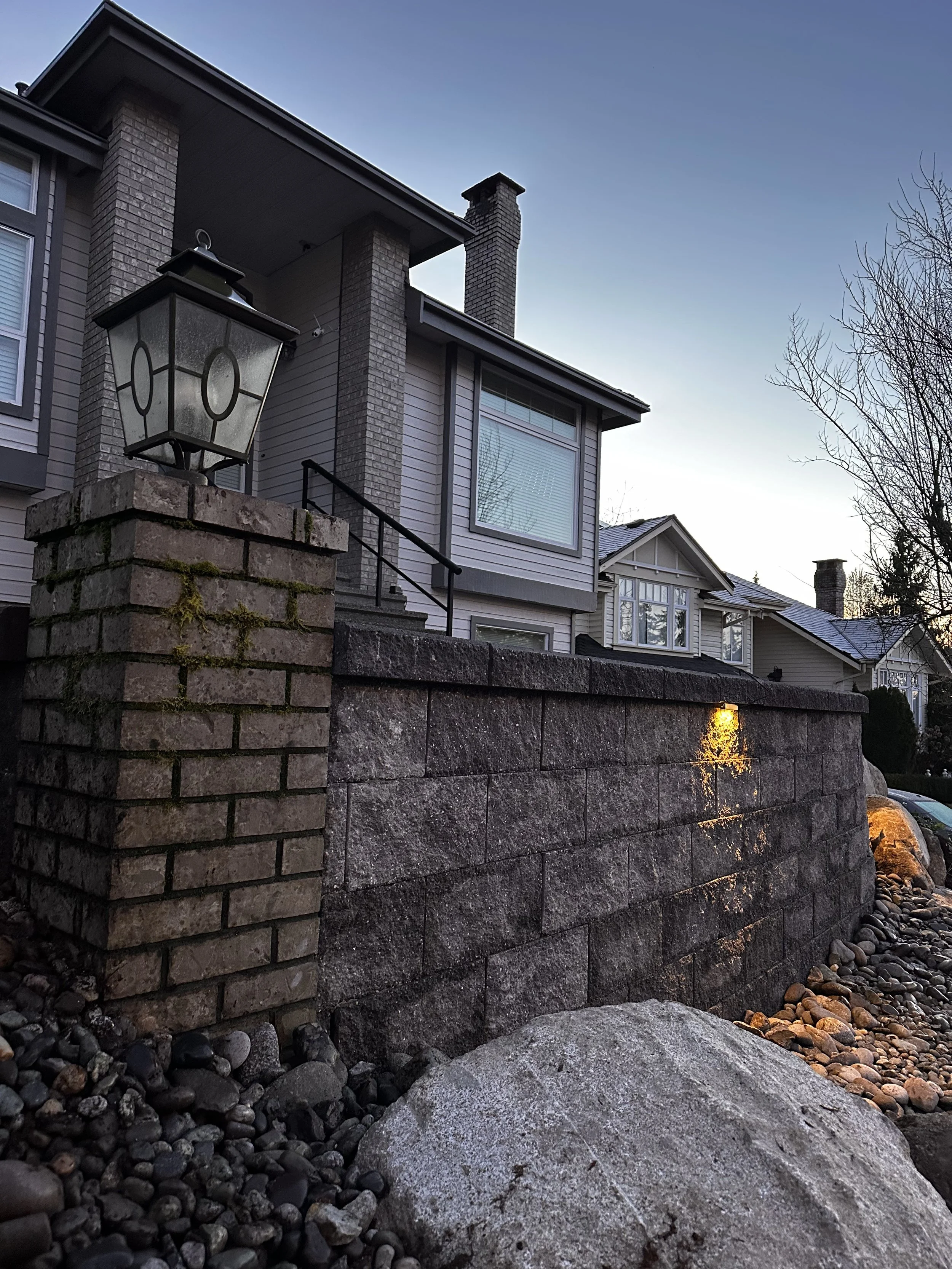 A brick and stone wall with an outdoor lamp on a pillar, in front of a modern house with large windows and a chimney, during dusk.