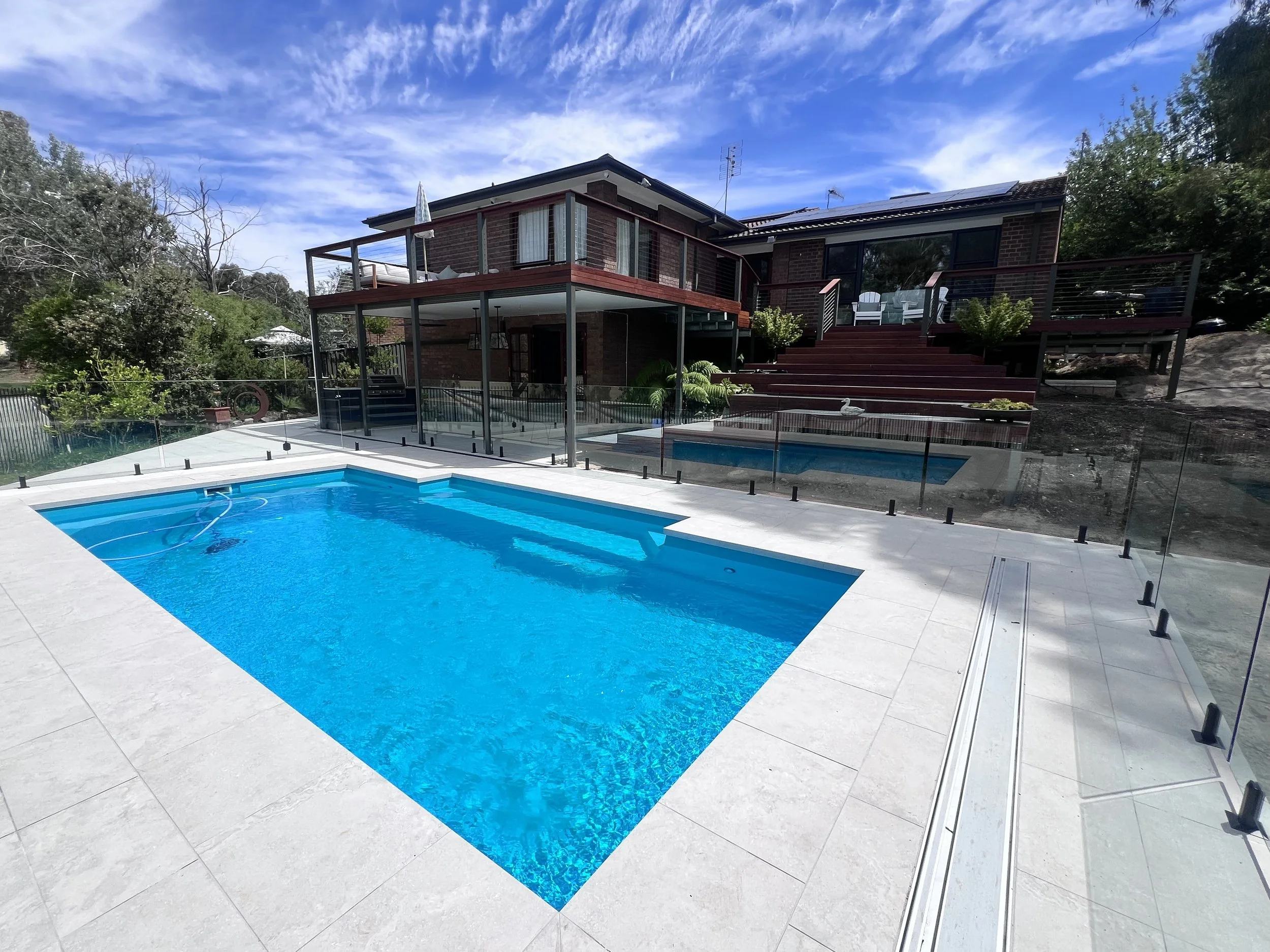 View of a backyard with a swimming pool, deck, and a two-story brick house with large windows and a balcony, surrounded by trees and a partly cloudy blue sky.