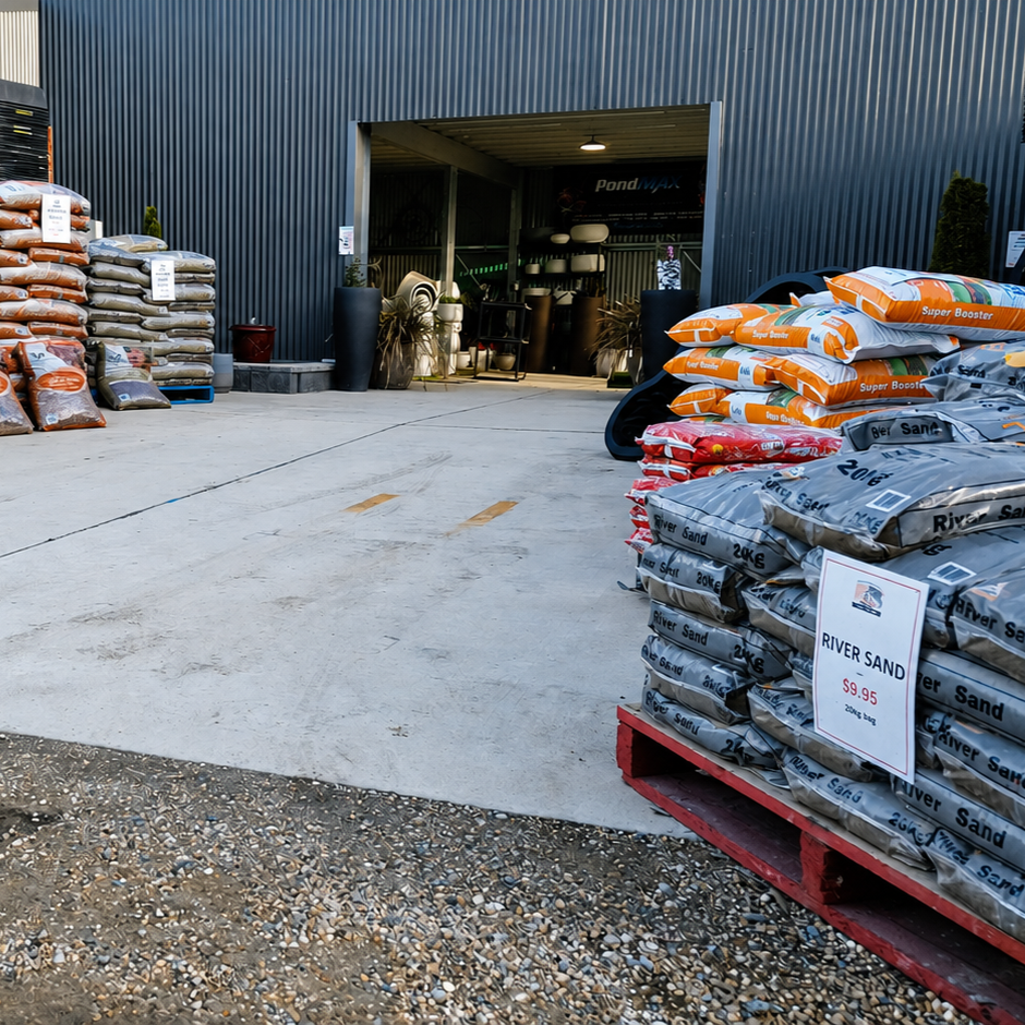 Exterior of a garden center with bags of river sand on a red pallet in the foreground, and stacked fertilizer bags to the left. The entrance to the store has various gardening supplies visible inside.