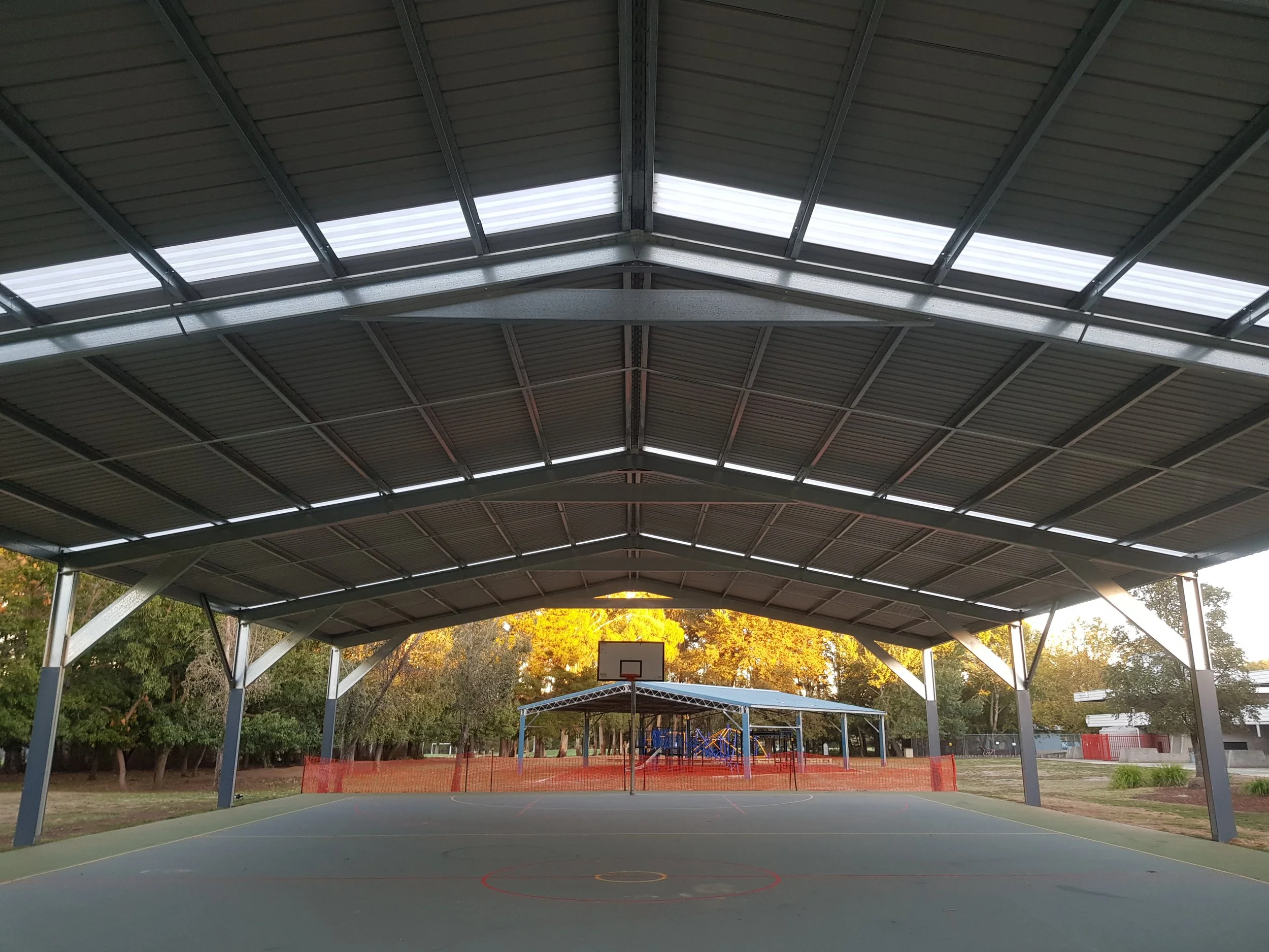 An outdoor basketball court with a large metal roof overhead, surrounded by trees and a fence, with playground equipment in the background.