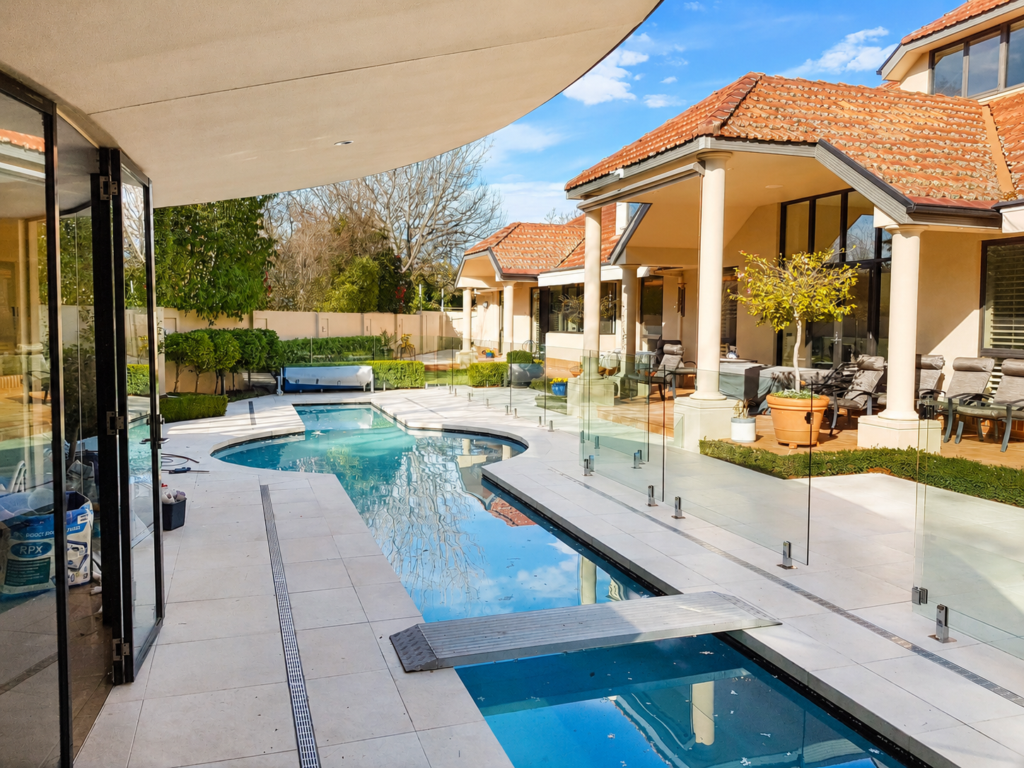 Modern backyard with a swimming pool, surrounded by a glass fence, patio furniture, potted plants, and neighboring houses with red-tiled roofs under a blue sky.