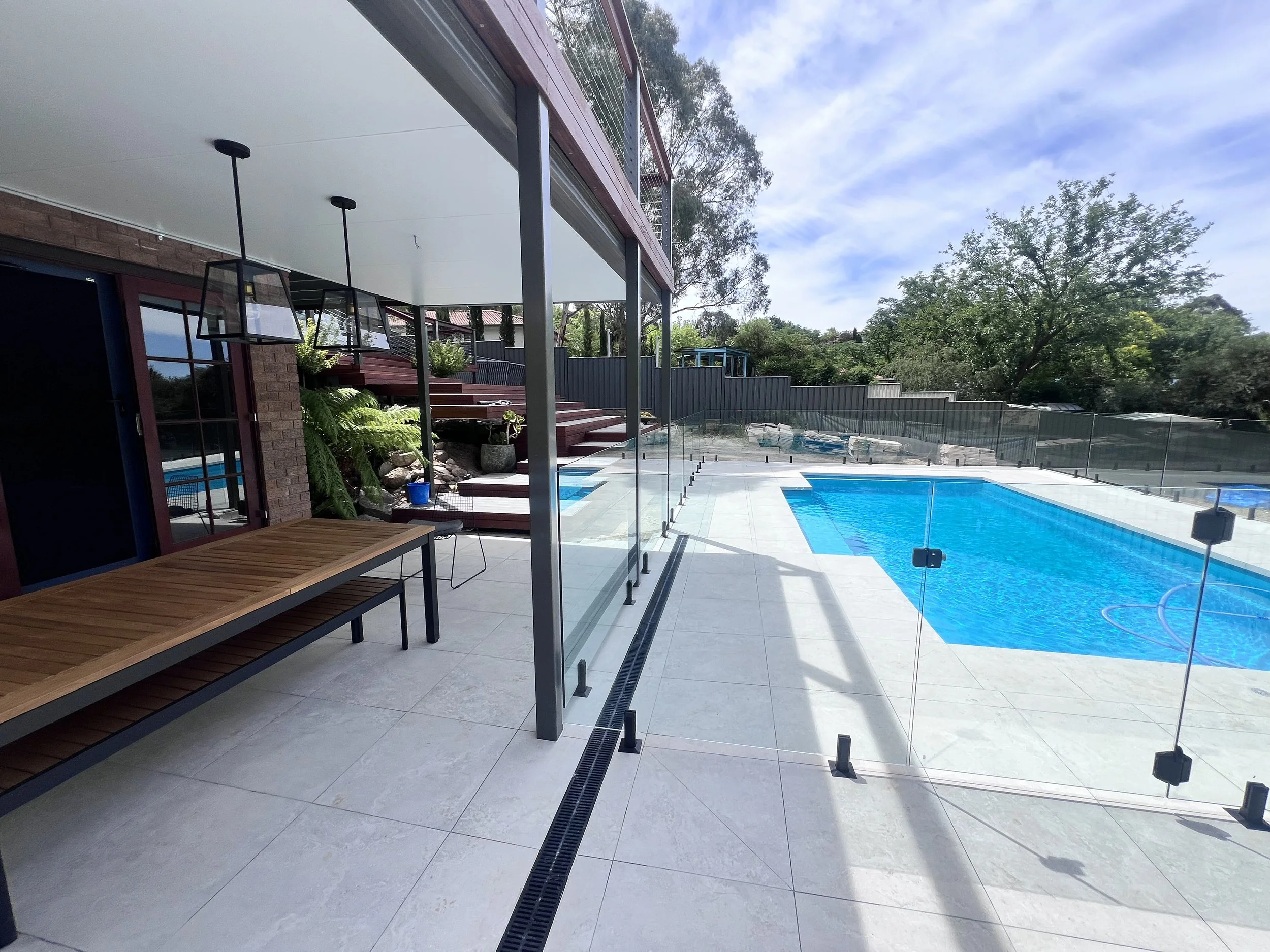 An outdoor swimming pool area with a glass fence, surrounded by trees and a partly cloudy sky.
