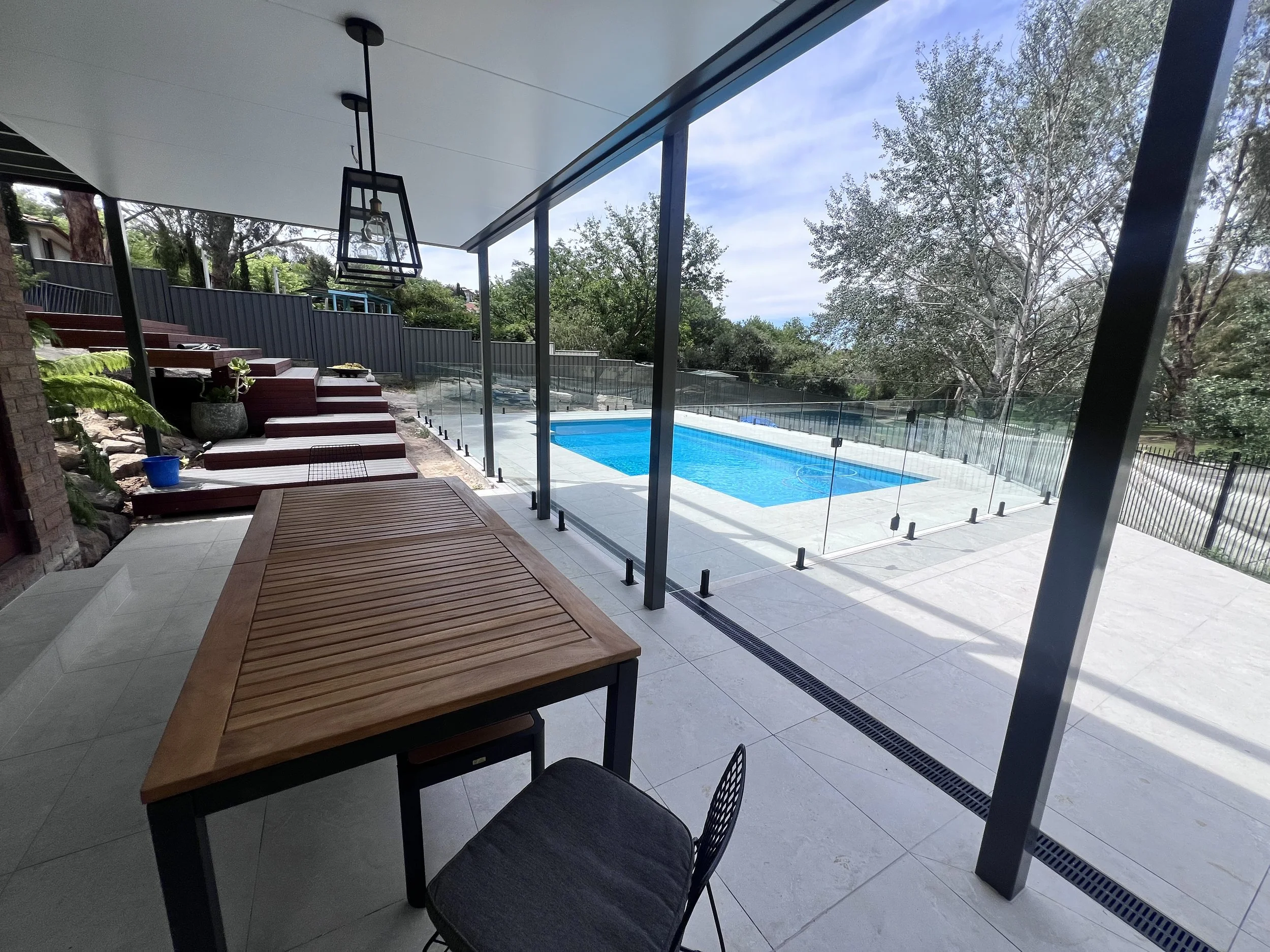 Modern outdoor patio with a glass-enclosed swimming pool, wooden deck, and a table and chairs. Trees and a fence are in the background under a partly cloudy sky.