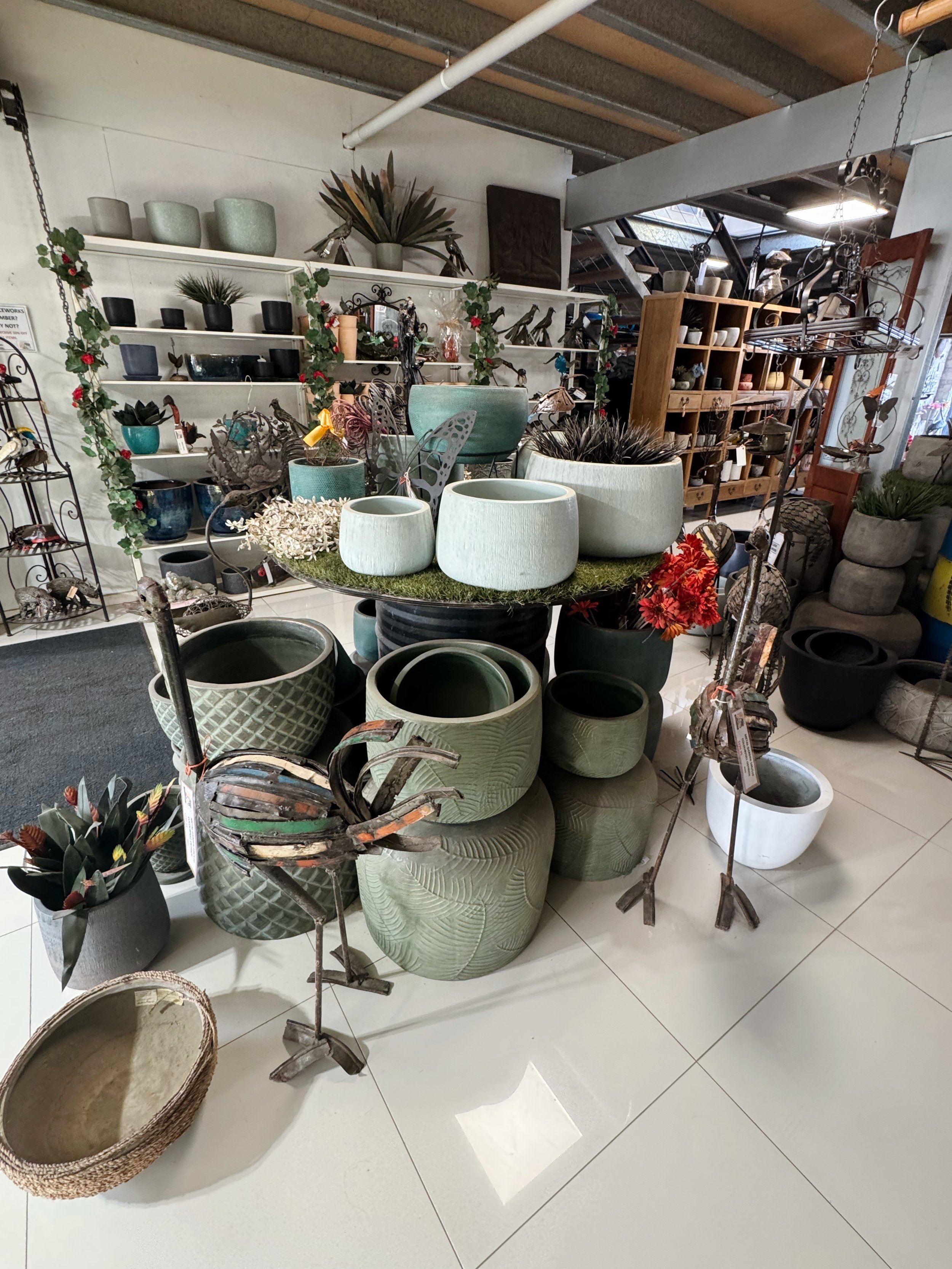 Display of various ceramic pots and planters at a store, with some decorative metal and artificial plants, shelves, and wooden furniture in the background.