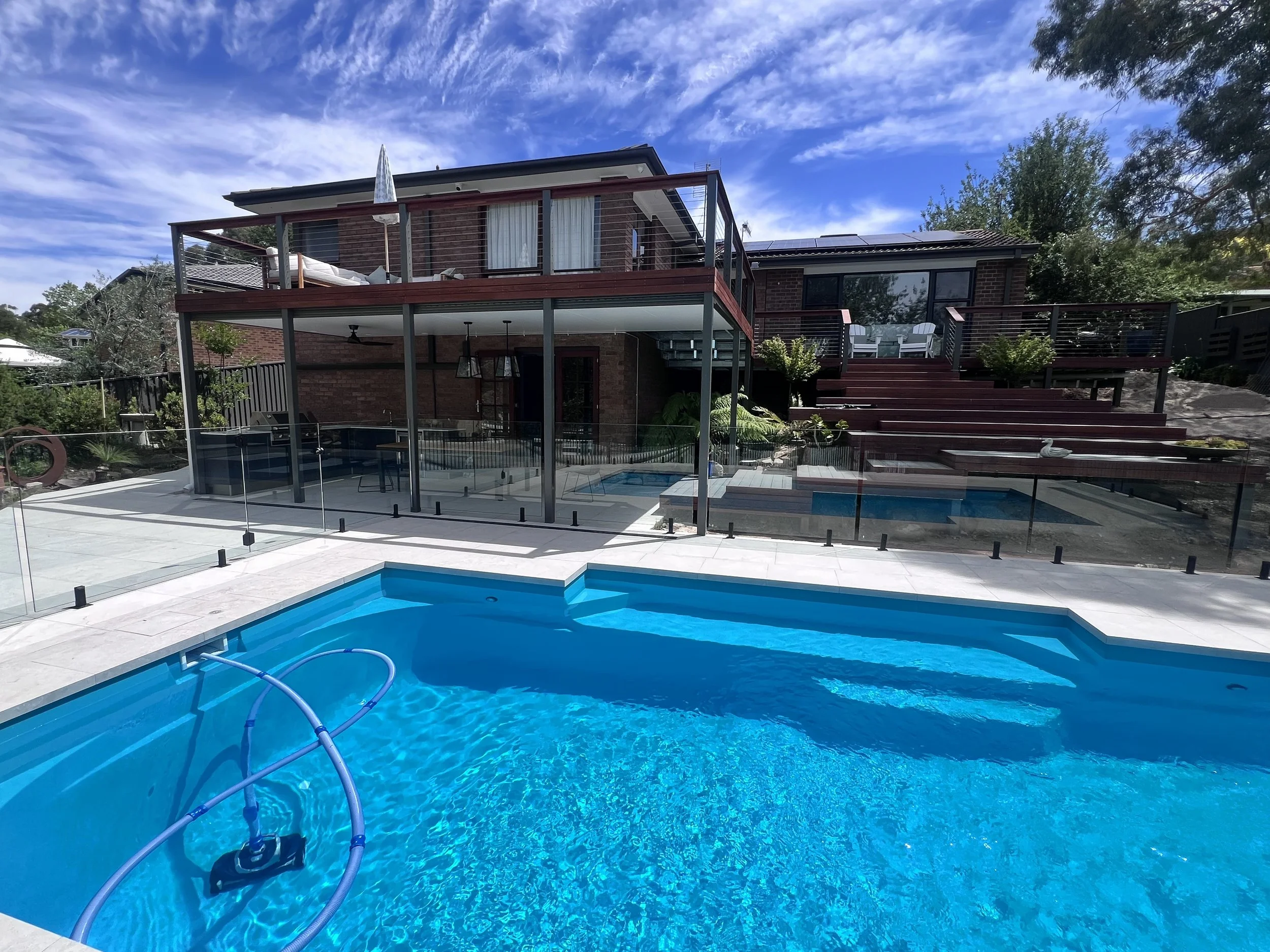 A backyard swimming pool with a safety fence, featuring blue water, pool cleaning equipment, and surrounding patio area. In the background, there is a two-story brick house with an upper deck and outdoor furniture, under a partly cloudy sky.