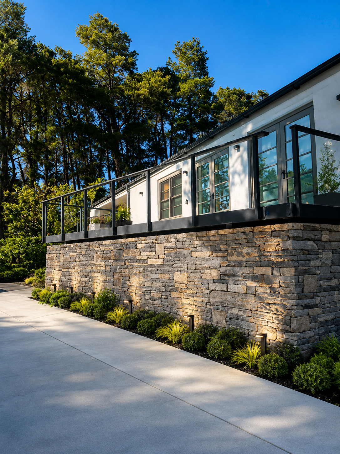 Modern house with a stone foundation, large windows, and a black metal railing on a balcony, surrounded by greenery and trees under a bright blue sky.