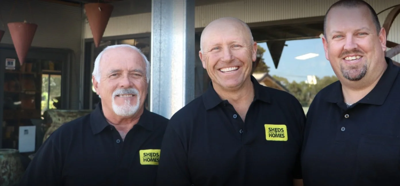 Three men wearing black shirts with yellow patches that say "Sheds & Homes" standing outdoors and smiling.