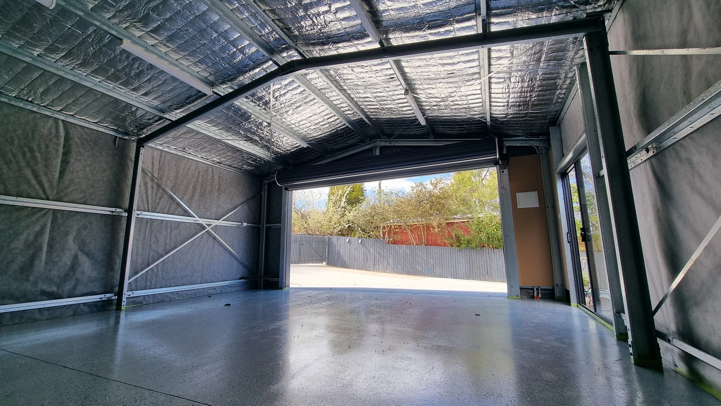Interior view of a garage under construction with exposed metal framing and insulation, an open garage door revealing a backyard with trees and a fence, and some construction materials on the floor.
