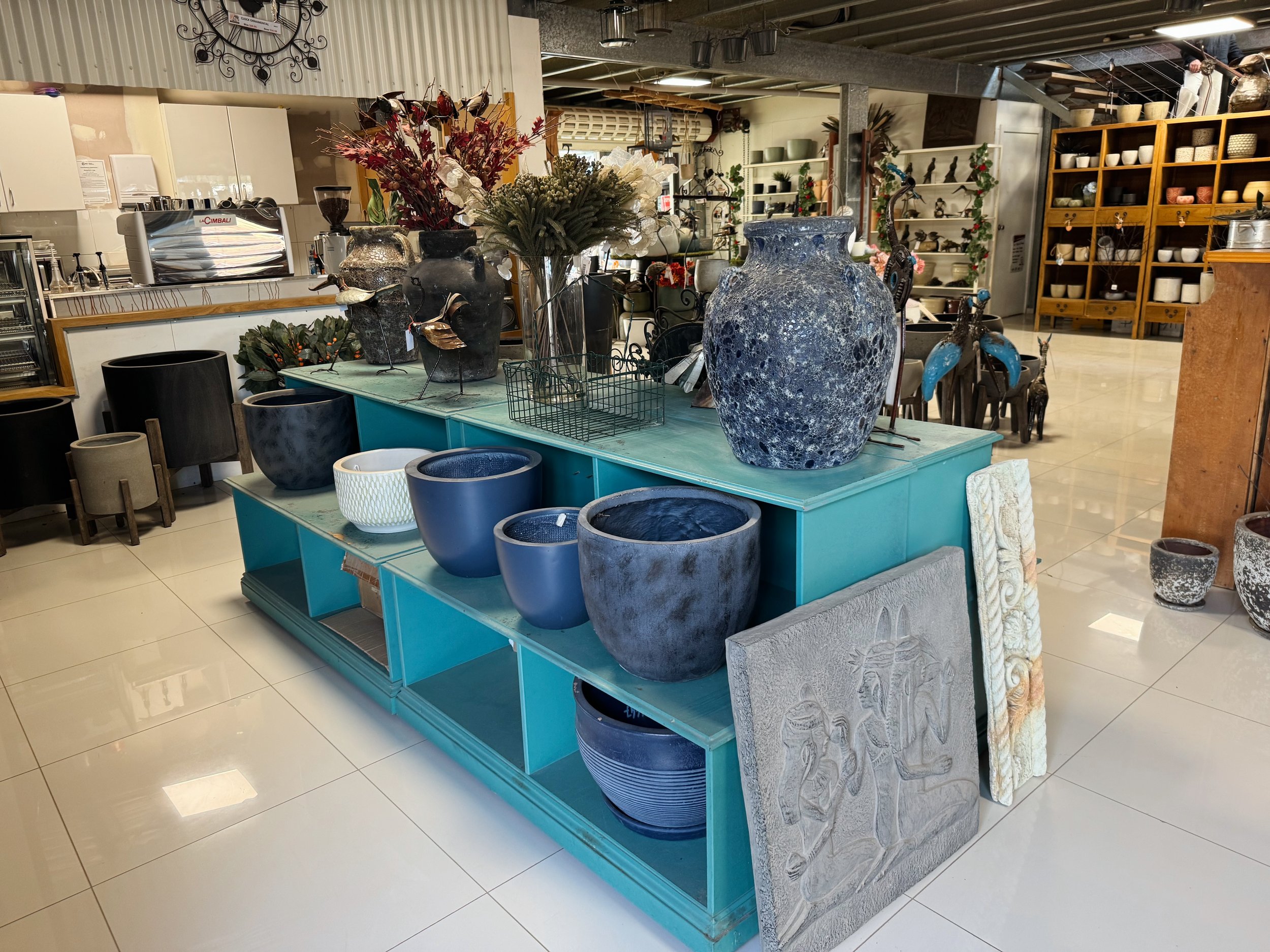 A store interior with various ceramic and decorative vases, pots, and sculptures on display, including a large blue textured vase on a turquoise stand and a flower arrangement in a glass vase. In the background, there are shelves filled with bowls, s