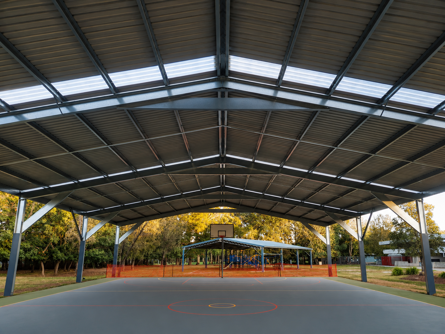 Empty outdoor basketball court with a roof structure, surrounded by trees and orange safety fencing.