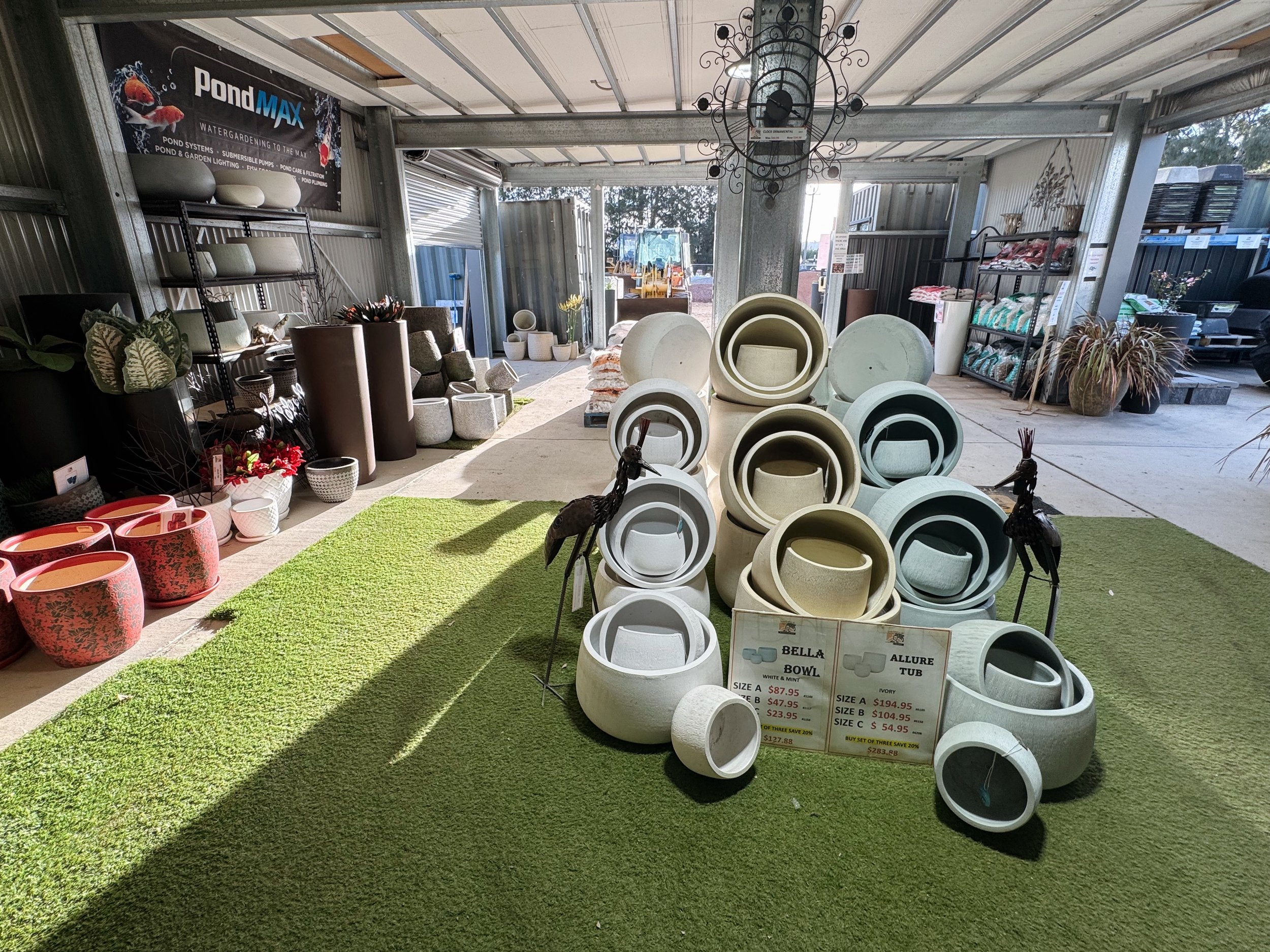 Interior view of a garden and outdoor decor shop with ceramic planters, statues, and garden ornaments displayed for sale. Sunlight filters through open garage doors, illuminating the space. Various pots and garden ornaments are arranged on shelves and on the floor, with a sign advertising prices for bowls and urns.