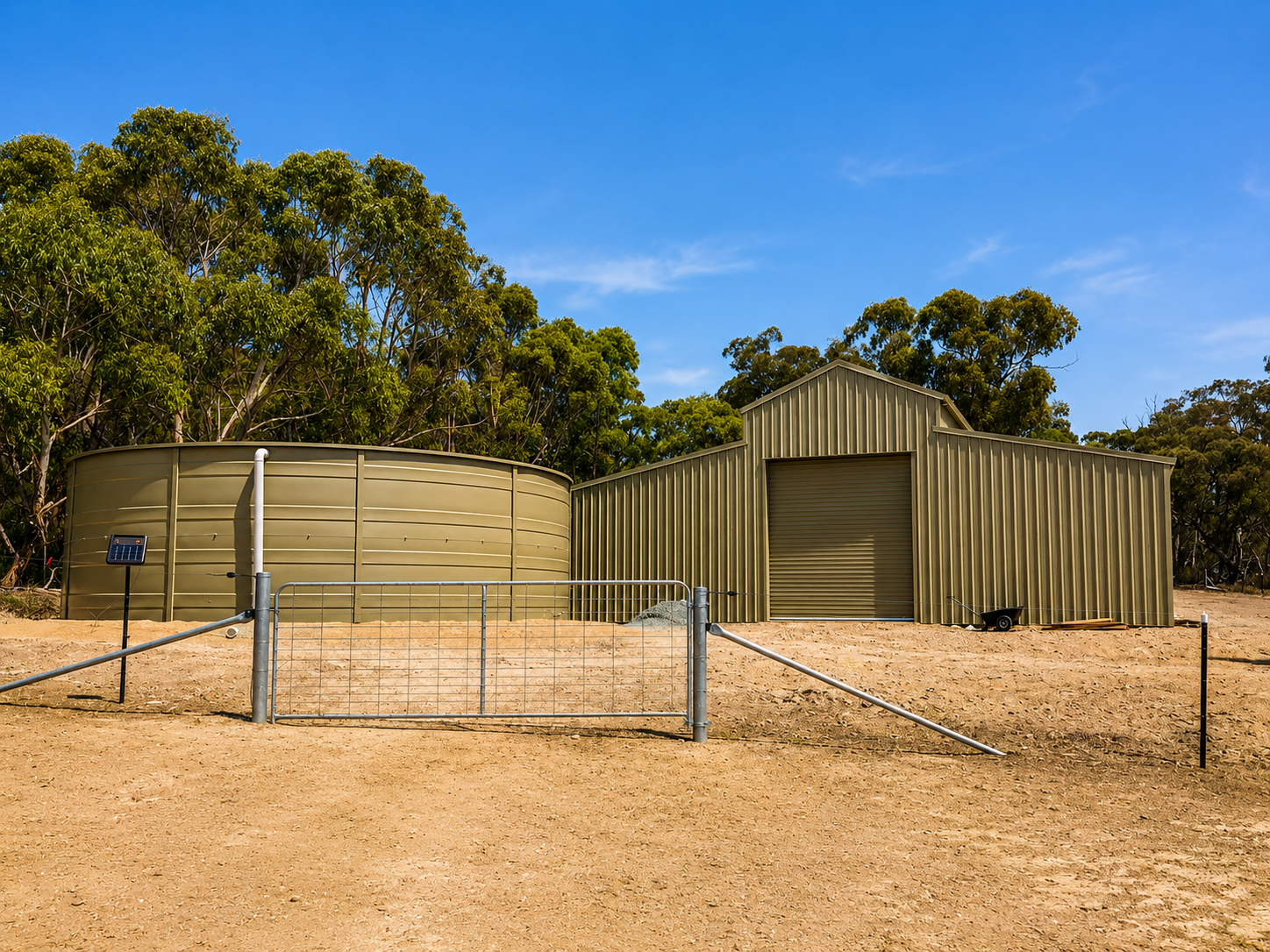 A rural scene with green metal storage shed and water tank on sandy ground, surrounded by trees and a blue sky.