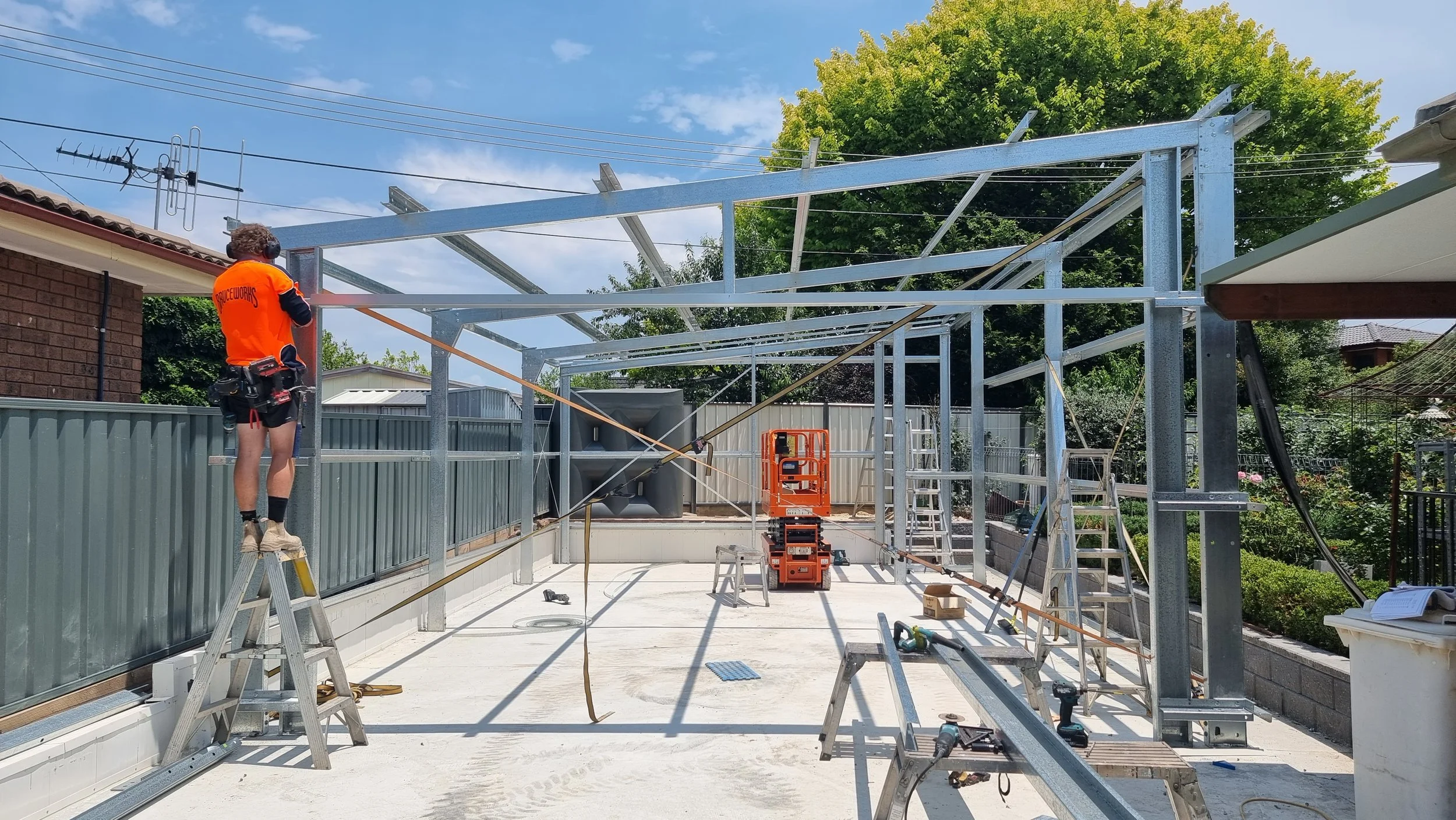 Construction worker in orange shirt and shorts installing metal frames for a building structure in a backyard, with ladders, tools, and safety equipment around.