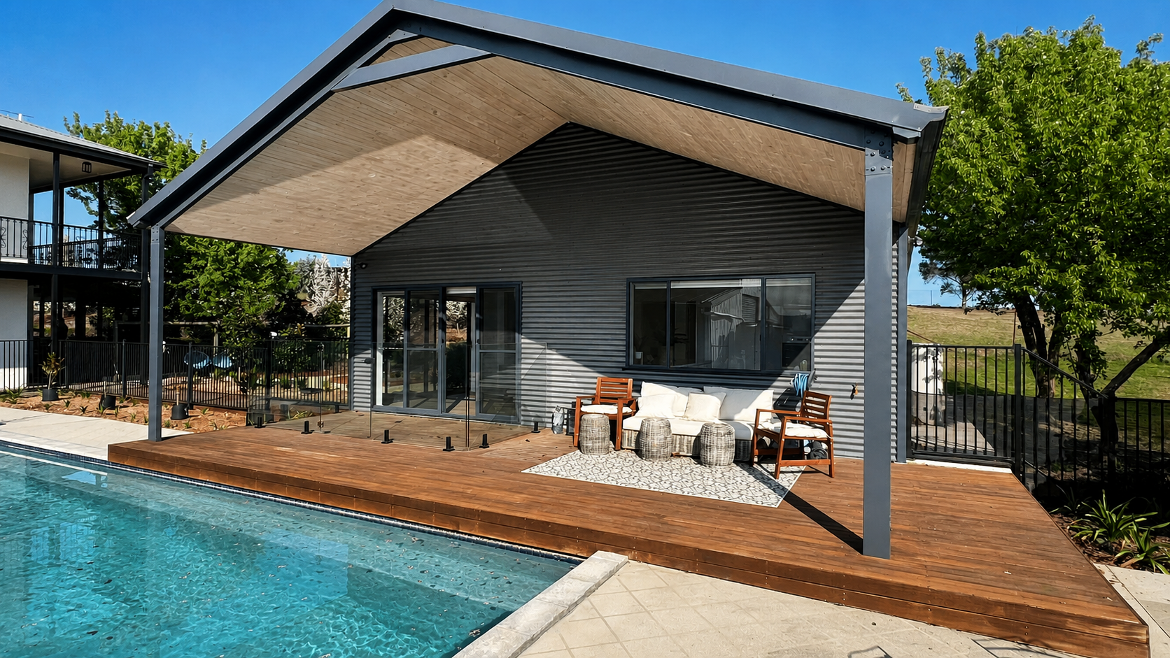 Modern house with a wooden deck and patio furniture beside a swimming pool, with a gray exterior, large umbrella, trees, and a clear blue sky.