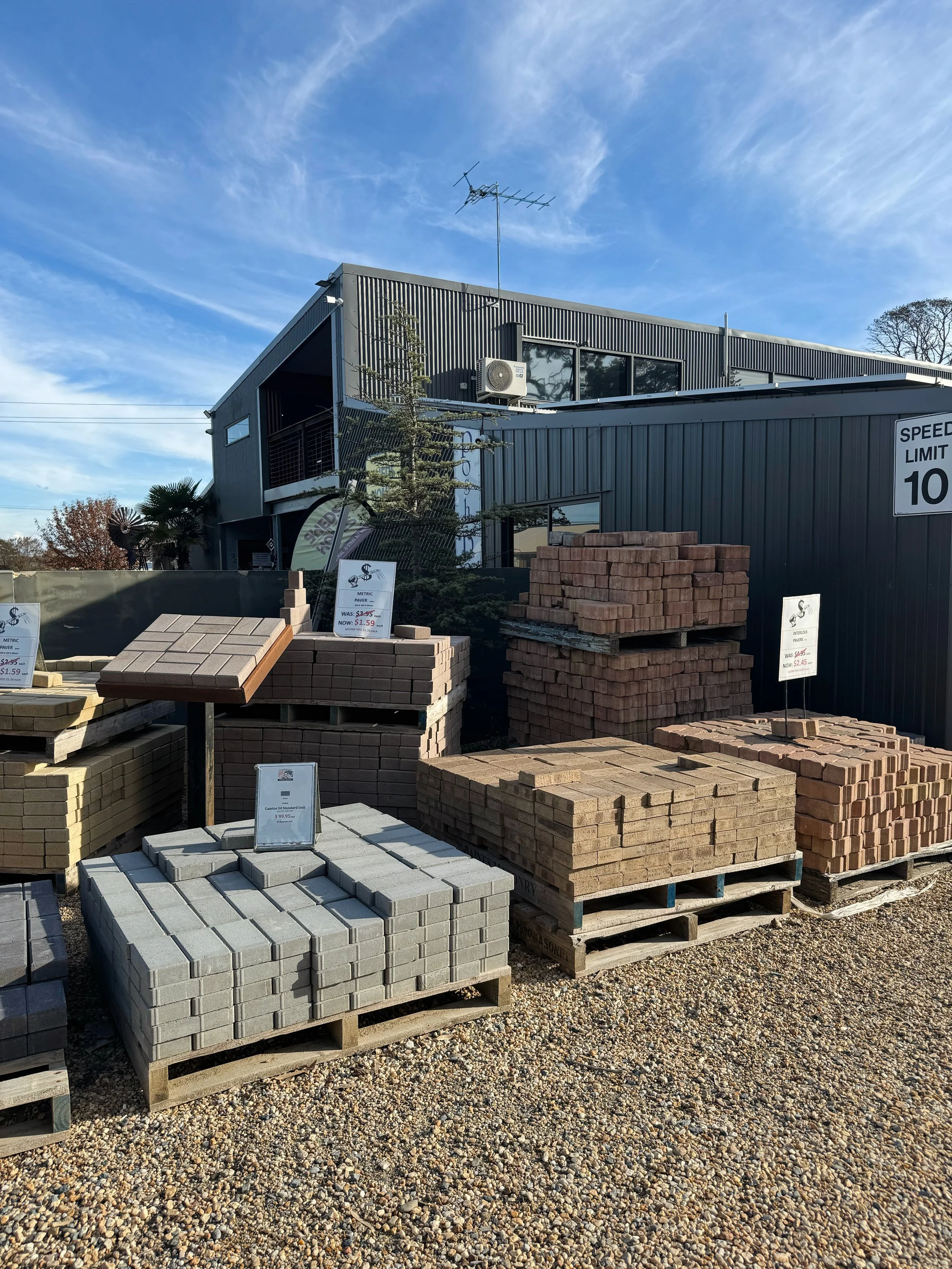 Display of various types of paving bricks and stones at an outdoor store with a building in the background, under a blue sky.