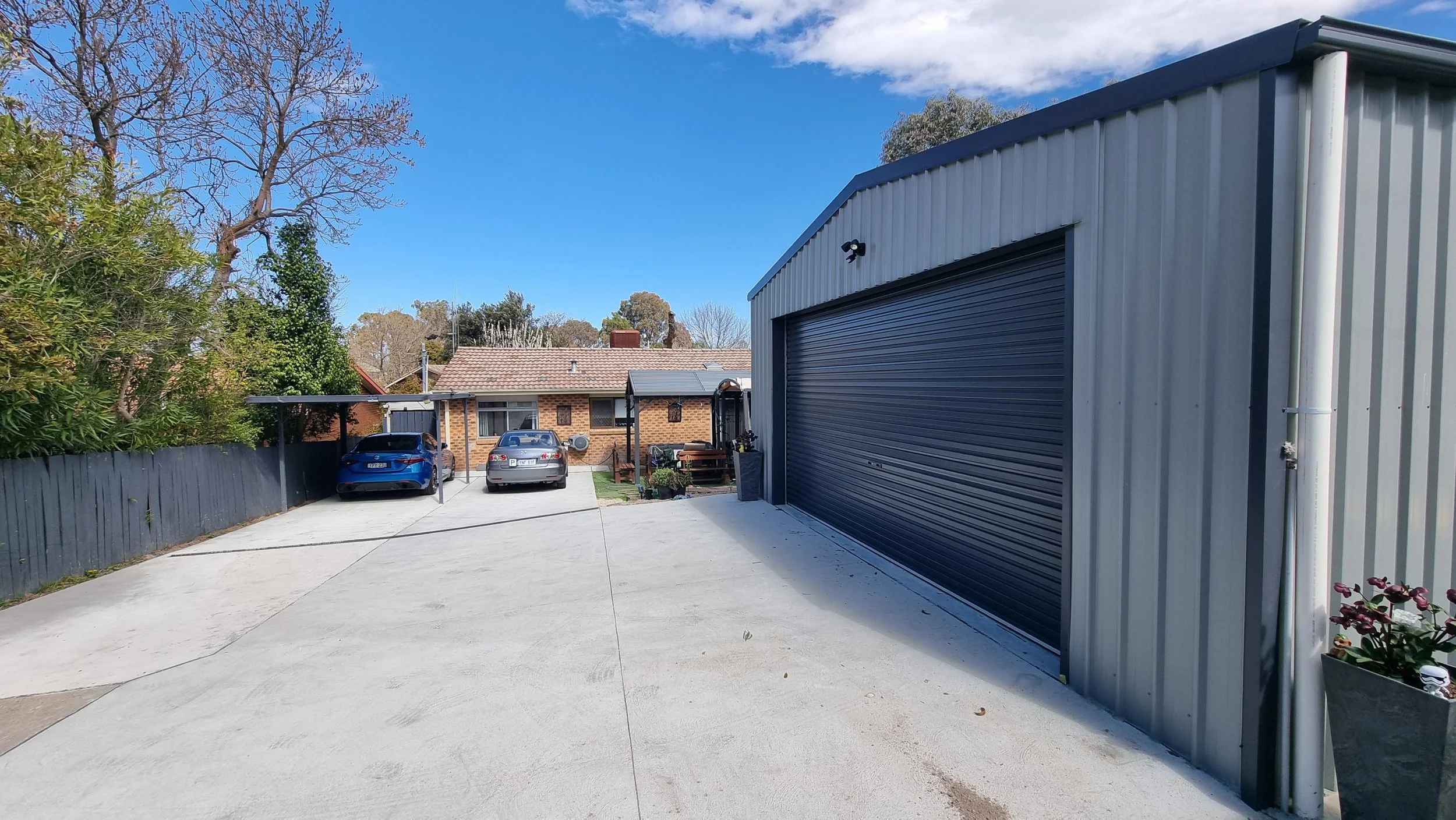 A concrete driveway in front of a metal garage with a dark roller door. Two cars are parked under a carport attached to a house with a brick wall. Trees with sparse leaves and a partly cloudy blue sky are visible in the background.