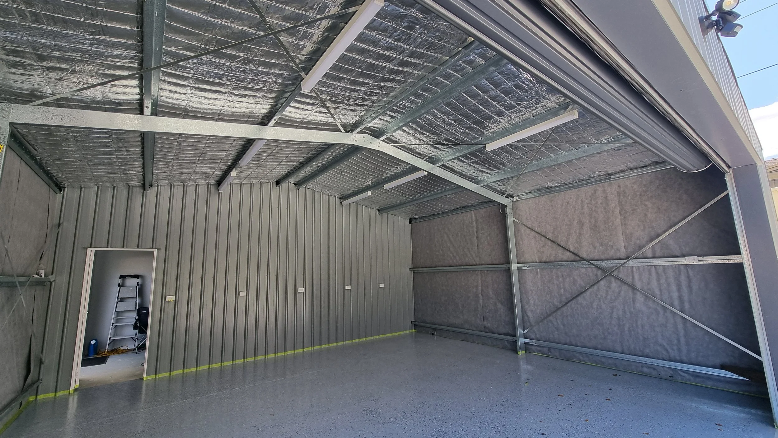 Interior of an unfinished metal garage or storage building with exposed insulation and framing, plywood flooring, a tiered shelf, and a ladder in a small room at the back.