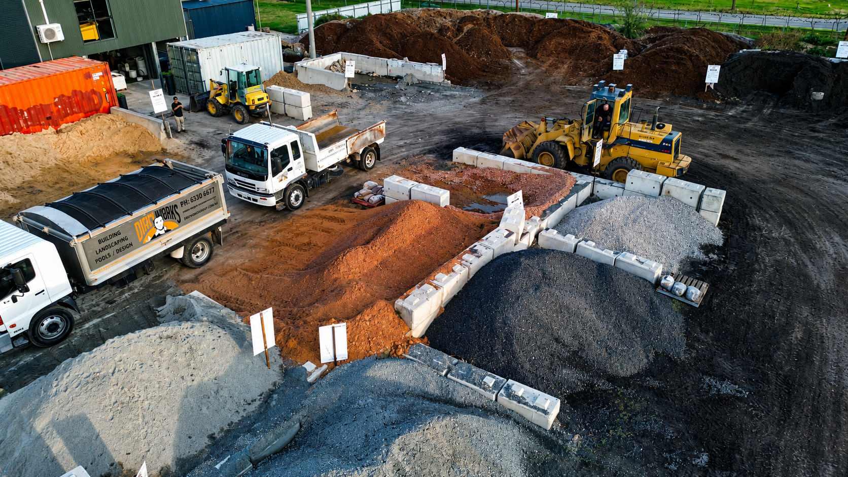 Construction site with piles of dirt, gravel, and sand. Several trucks, including a dump truck, a front loader, and a small yellow bulldozer. White barriers and signs mark different sections of the site. Gravel and dirt are being moved and organized.