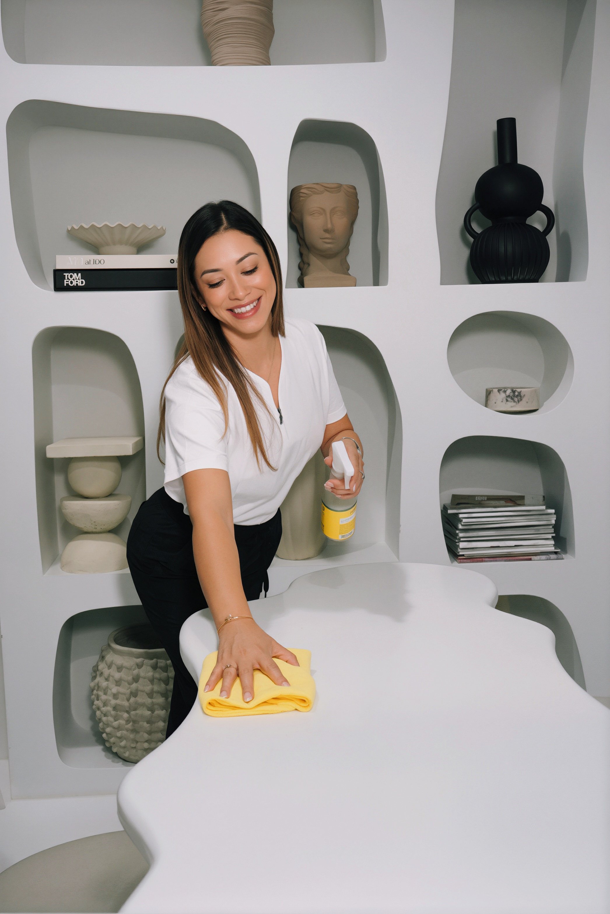 Woman cleaning a white, modern desk with a yellow cloth, smiling and looking down, in a minimalist office decorated with white and beige ceramics and books.