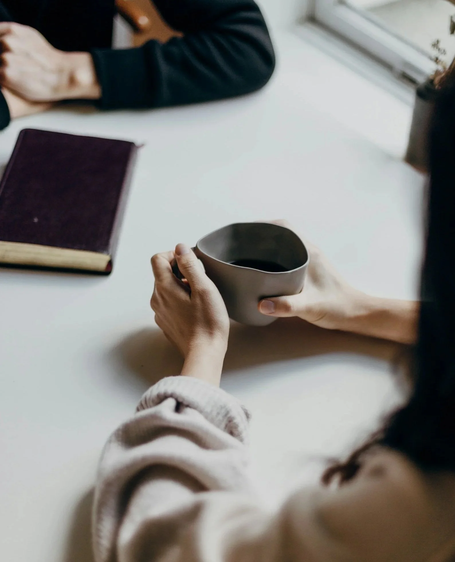 People sitting around a table, one person is holding a coffee mug and there is a closed book with a dark cover on the table.