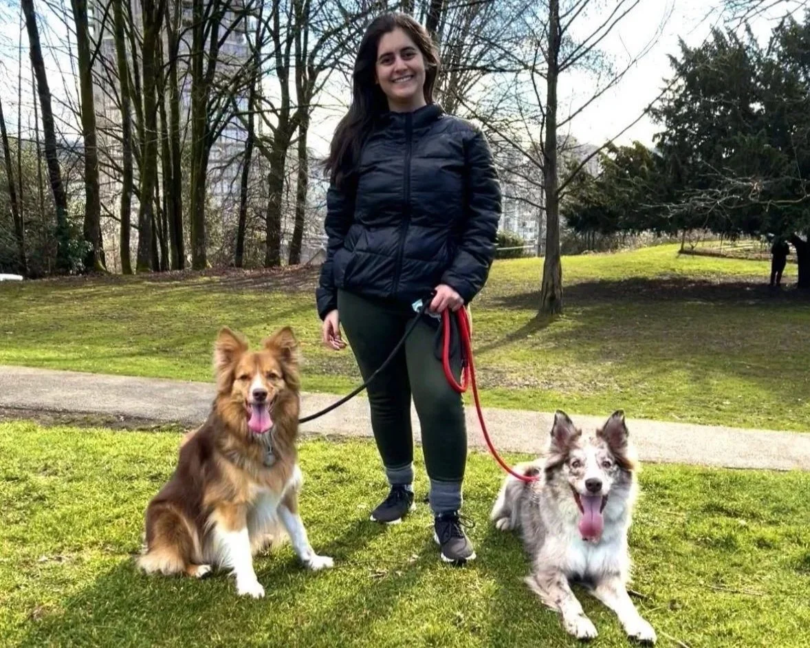 Mulher sorrindo segurando as coleiras de dois cães, um sentado ao lado esquerdo com pelagem marrom e branco, e outro deitado ao lado direito com pelagem mesclada de cinza, branco e preto, em um parque com árvores e grama.