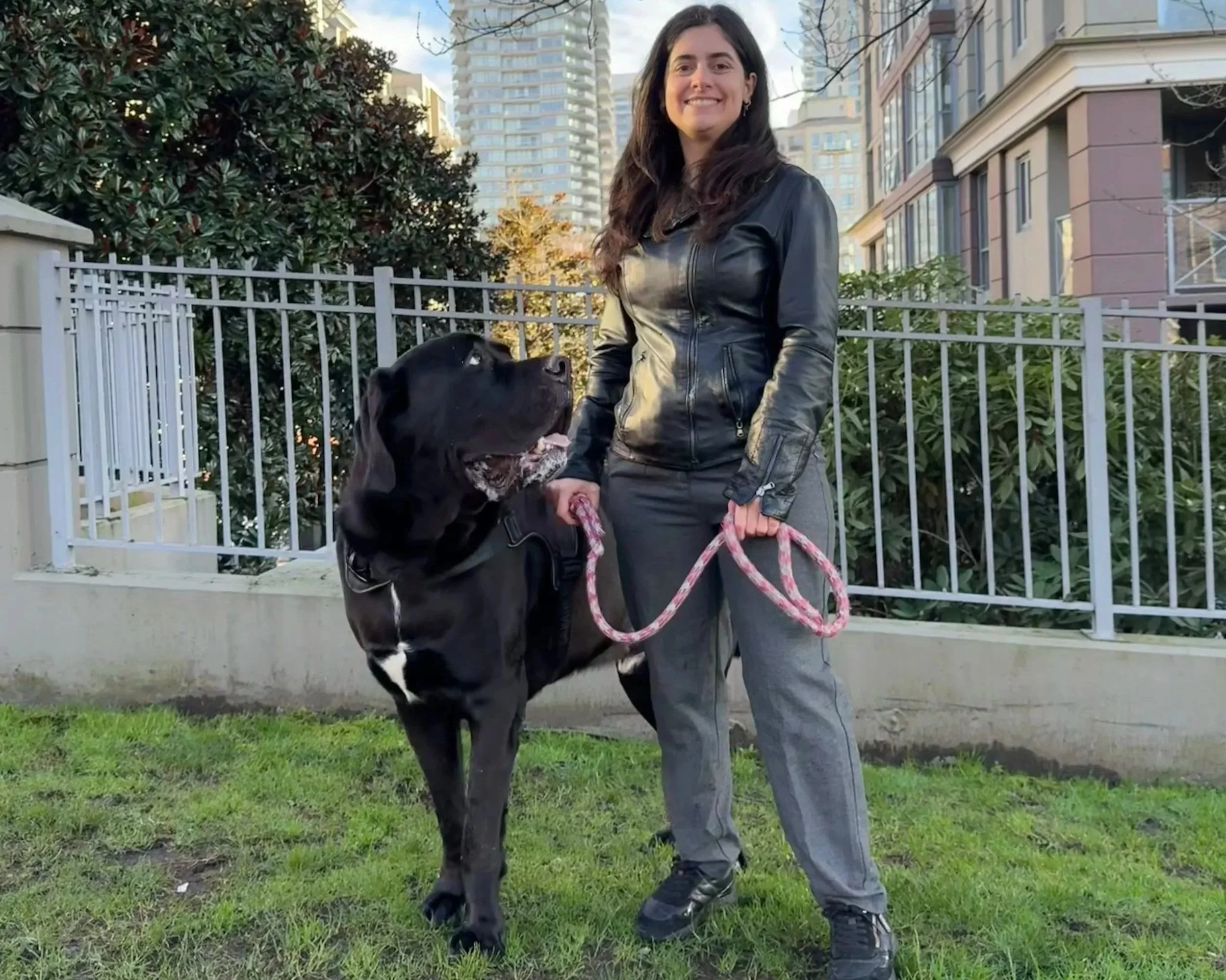 Pessoa sorridente de cabelos longos usando jaqueta de couro escura e calça cinza escura segurando a guia de um cachorro pastor alemão preto com manchas brancas na cabeça e peito, em área urbana com prédios altos ao fundo e cerca branca ao redor.