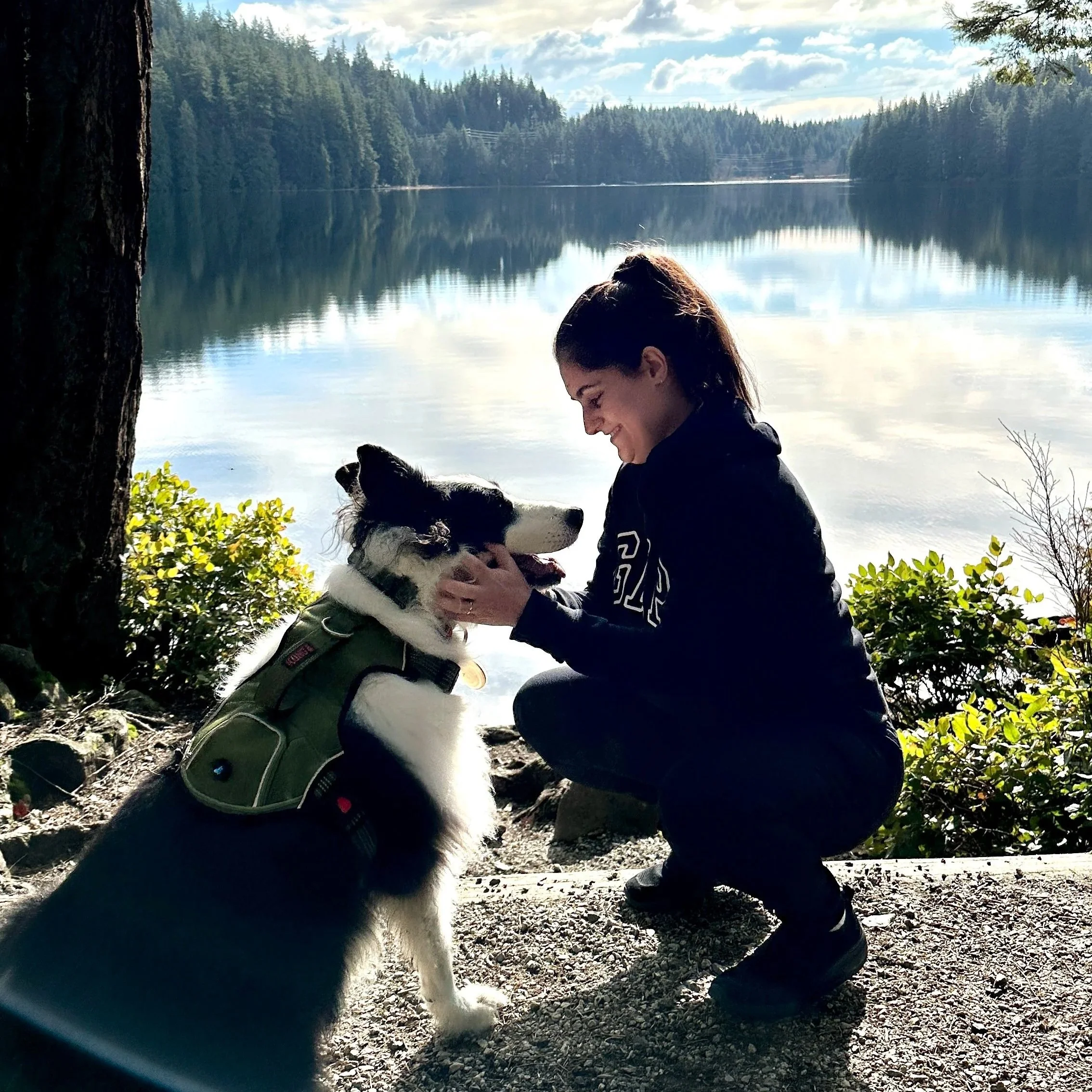 Pessoa sorridente com cabelo ao vento, segurando cabeça de cachorro de pelos brancos e pretos, próximo a uma árvore, com lago, árvores e céu com nuvens ao fundo.