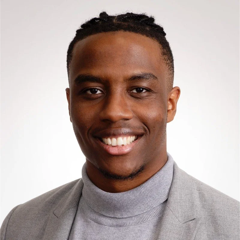 Headshot of a smiling African American man in a gray blazer and turtleneck, against a plain white background.