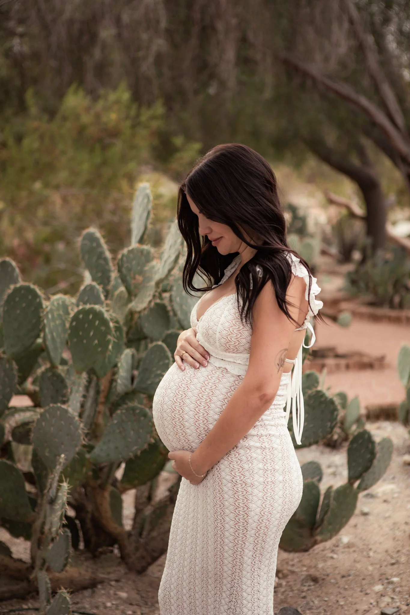 A pregnant woman with long dark hair standing outdoors among prickly pear cacti, wearing a white lace dress and looking down with her arms crossed on her belly.