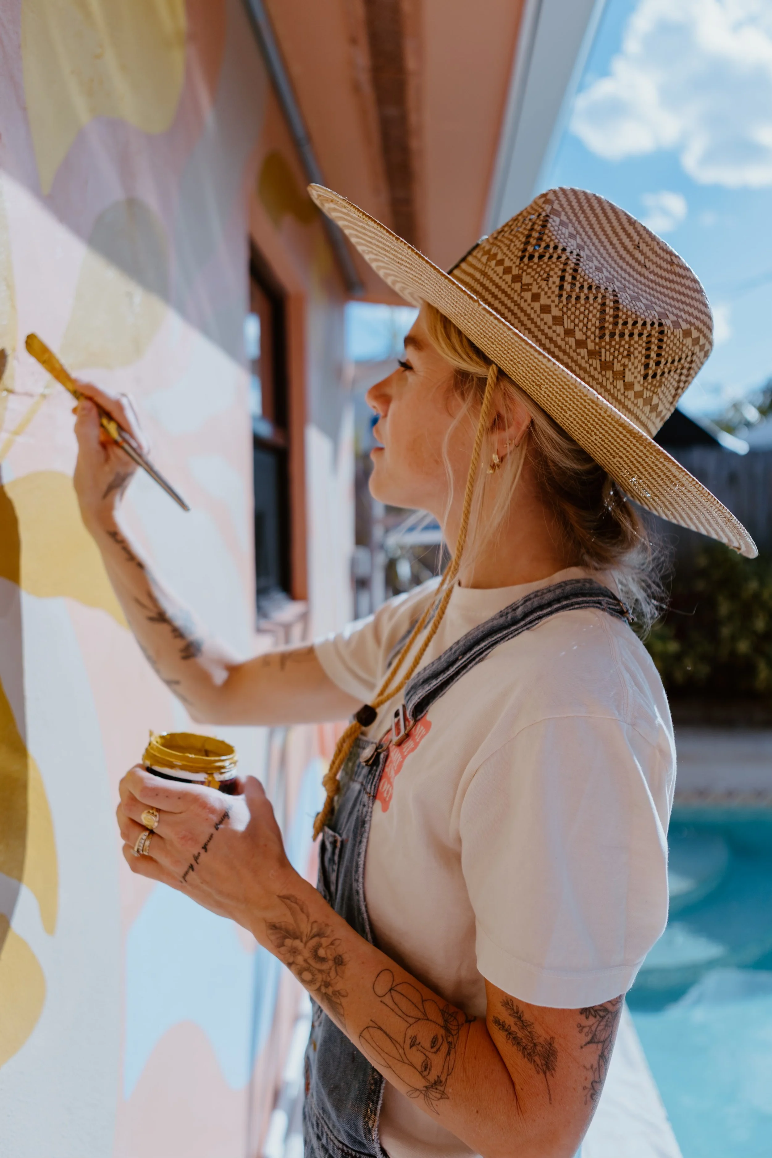 A woman wearing a wide-brimmed straw hat and a white t-shirt is painting a wall outdoors. She has tattoos on her arms and is holding a jar of yellow paint and a paintbrush. The sky is partly cloudy in the background.