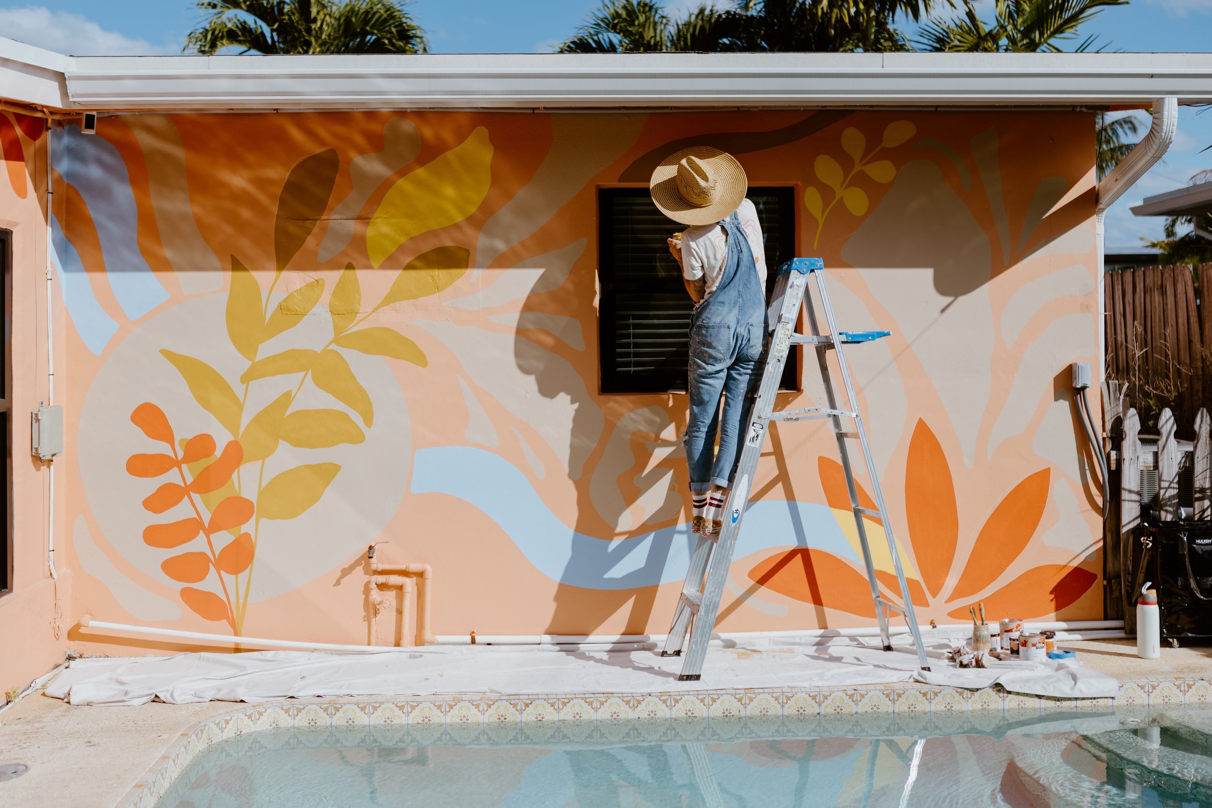 Person standing on a ladder painting a large mural on the exterior wall of a house. The mural has shades of orange, yellow, and pale blue with leafy patterns. The person wears a wide-brimmed straw hat, white shirt, and blue overalls. There is a pool in the foreground reflecting the scene, and painting supplies are on the ground.
