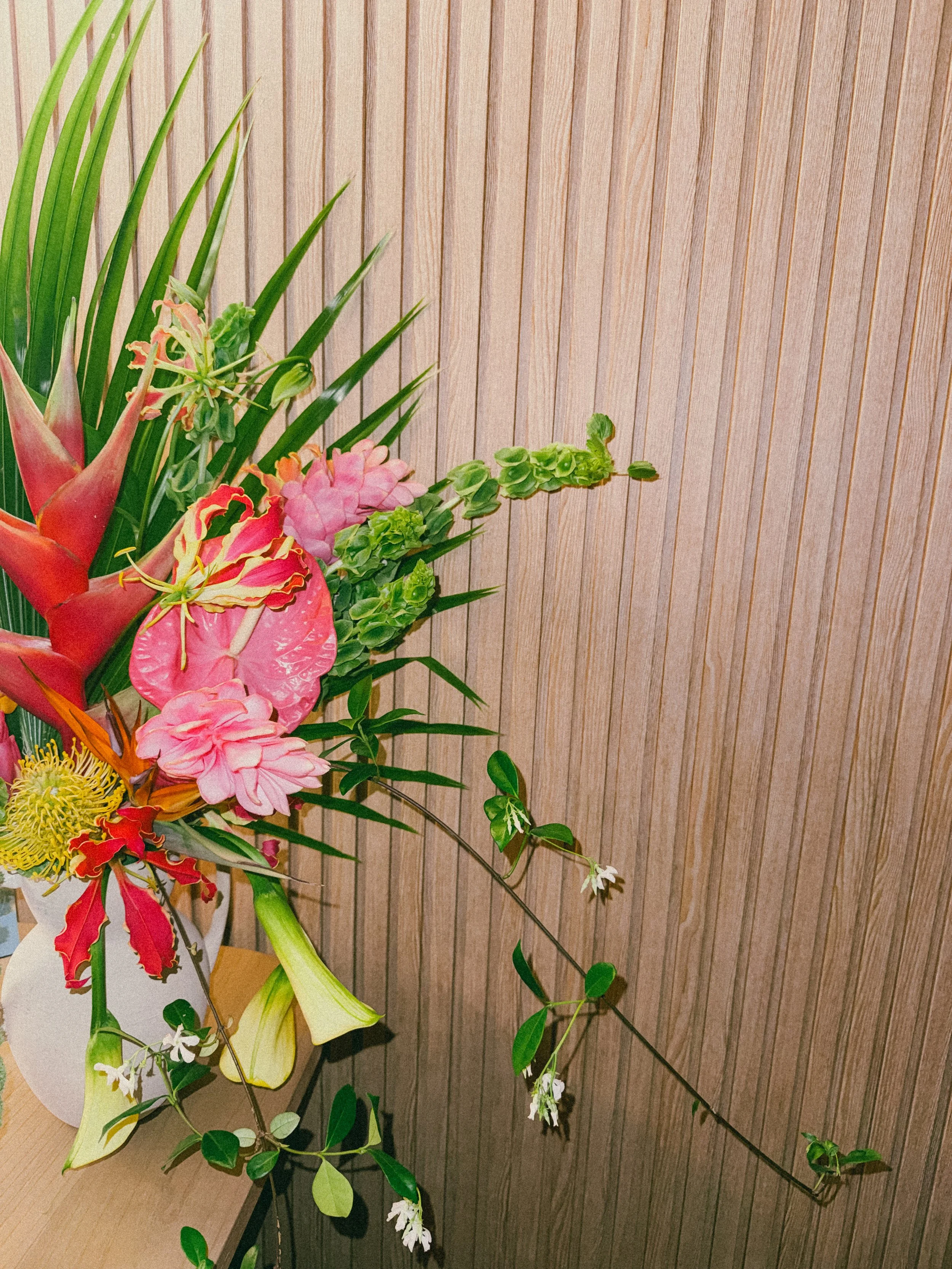 A colorful flower arrangement with pink, red, yellow, and white flowers, including lilies and other blossoms, against a wooden paneled background.