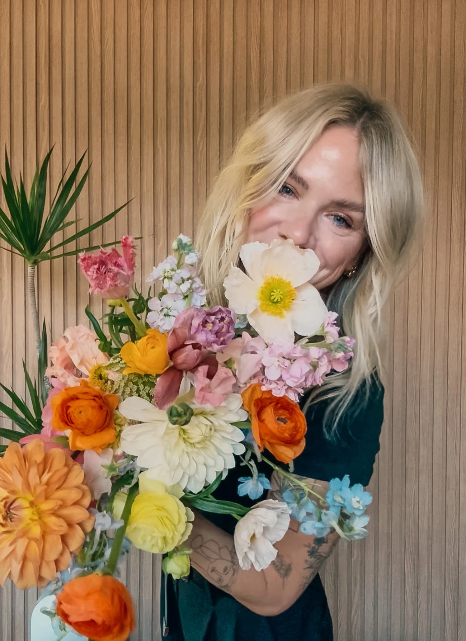 A woman with blonde hair holding a large bouquet of colorful flowers, including orange, pink, white, and yellow blooms, standing in front of a wooden paneled wall.
