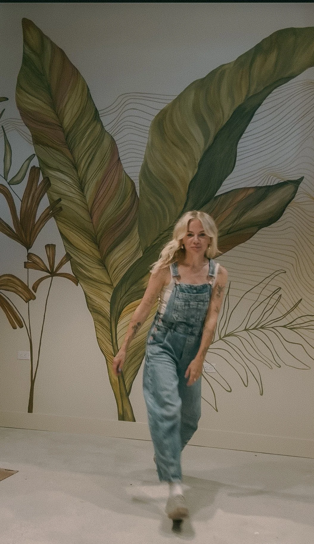 A woman with blonde hair, wearing overalls and white shoes, walking in front of a large wall mural of green and brown leaves.