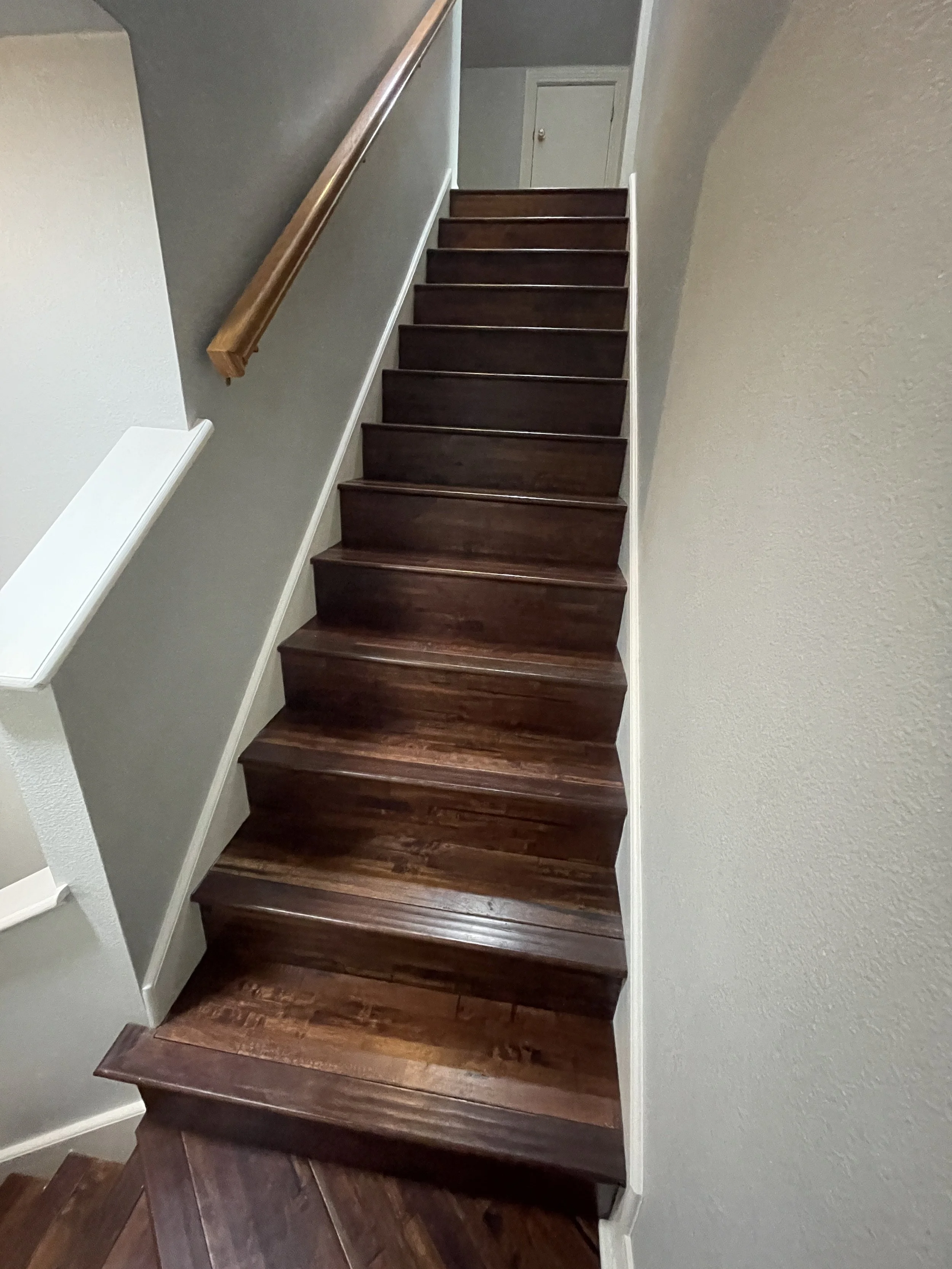A staircase with dark wooden steps, a wooden handrail on the left, white trim, and textured light-colored walls, leading to a closed door at the top.