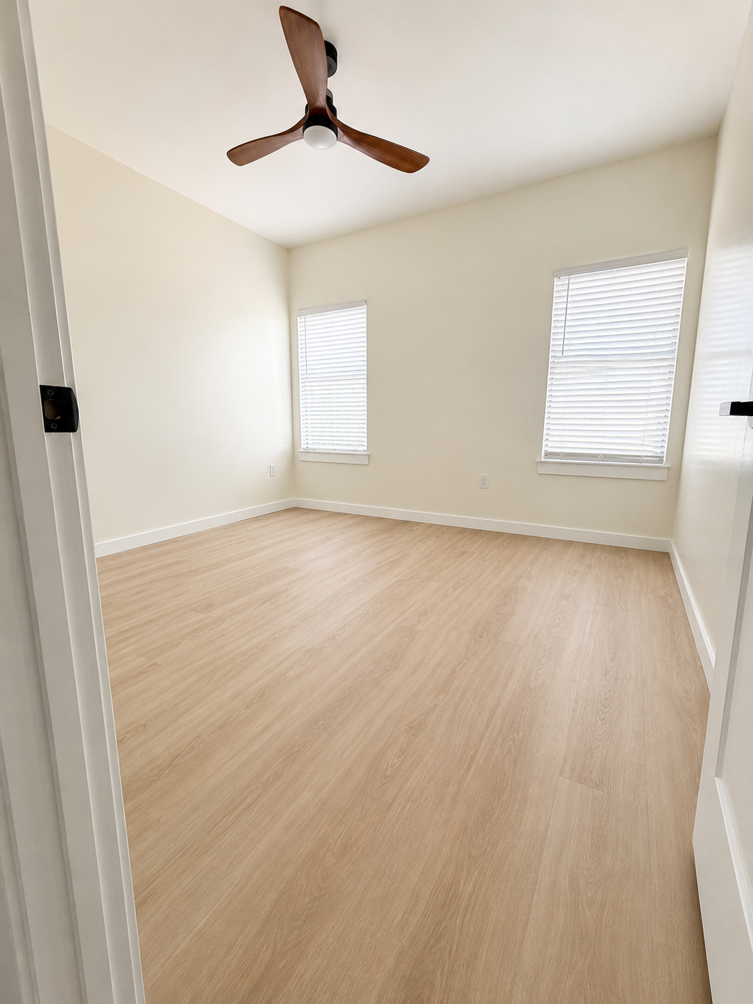 Empty room with light wood flooring, two windows with blinds, white walls, and a ceiling fan with wooden blades.