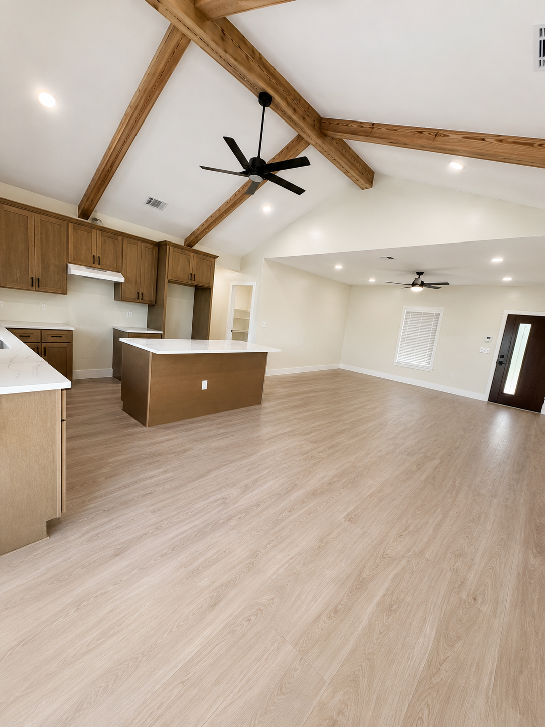 Open-concept living room and kitchen with vaulted ceiling, wooden beams, ceiling fans, light-colored hardwood flooring, white walls, dark front door, and window with blinds.