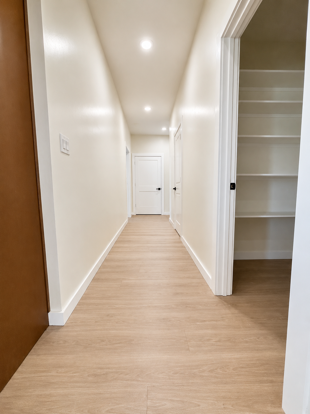 A hallway with beige walls and light wood flooring, featuring white doors and an open closet with built-in shelves.