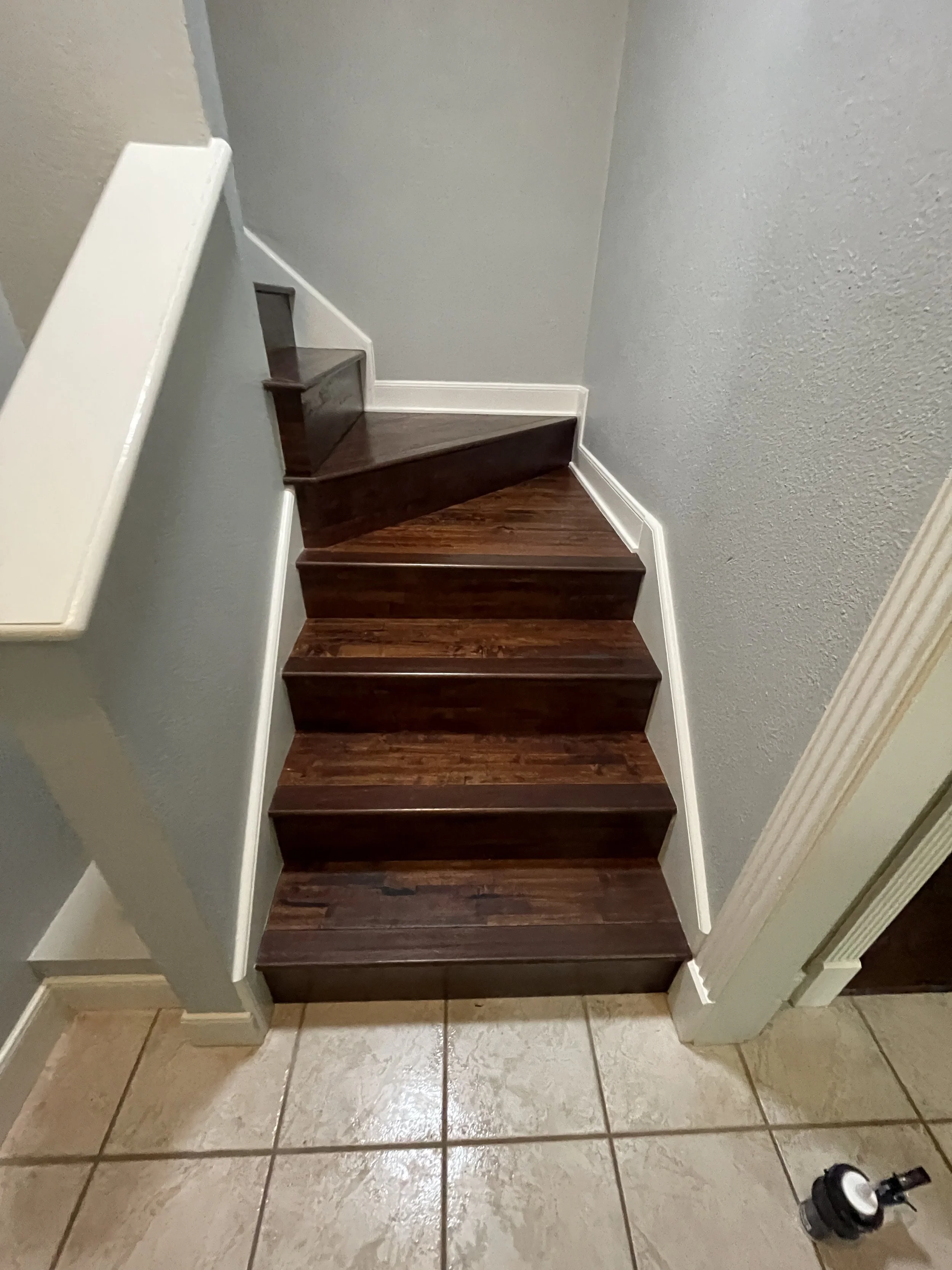 Interior view of a staircase with wooden steps and white trim, with beige tile flooring at the bottom. A black object, possibly a small vacuum or cleaning device, is on the floor near the bottom right corner.