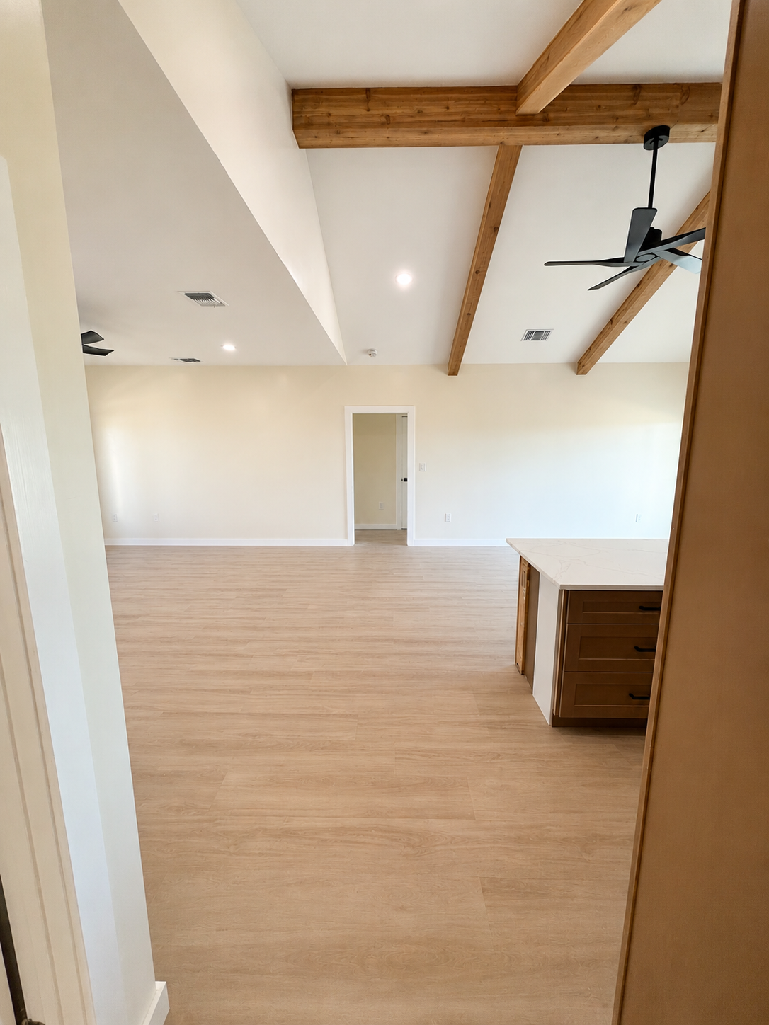Empty room with vaulted ceiling, wooden beams, ceiling fans, and light-colored flooring.