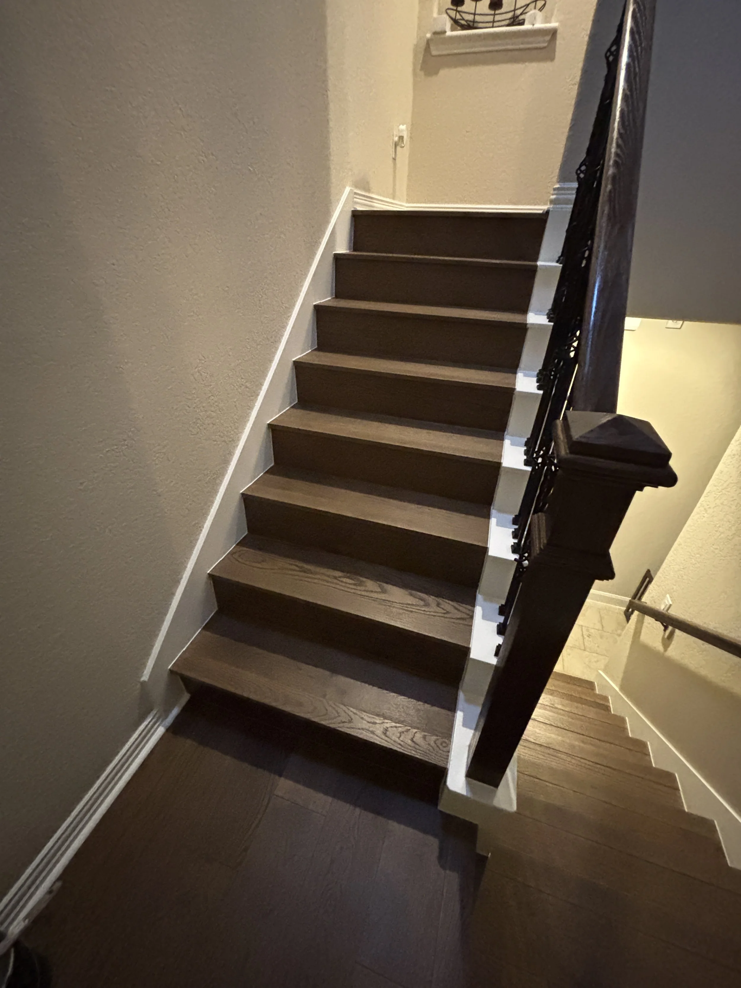 Interior view of a staircase with dark brown wooden steps and black decorative iron railing, beige walls, and a small window at the top of the stairs.