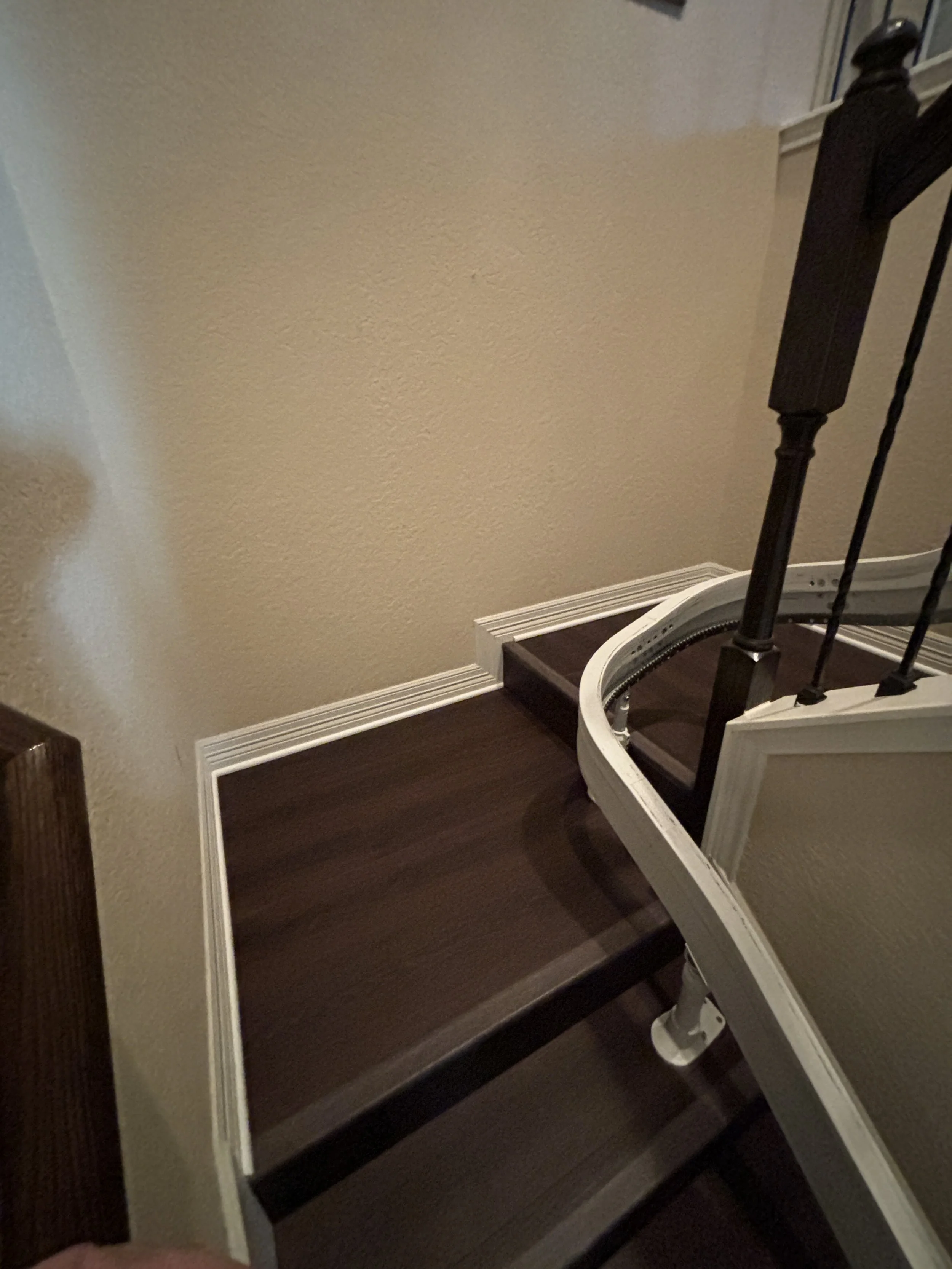 Inside view of a staircase with dark wooden steps, white trim, and a beige wall. Part of a black stair railing is visible on the right.