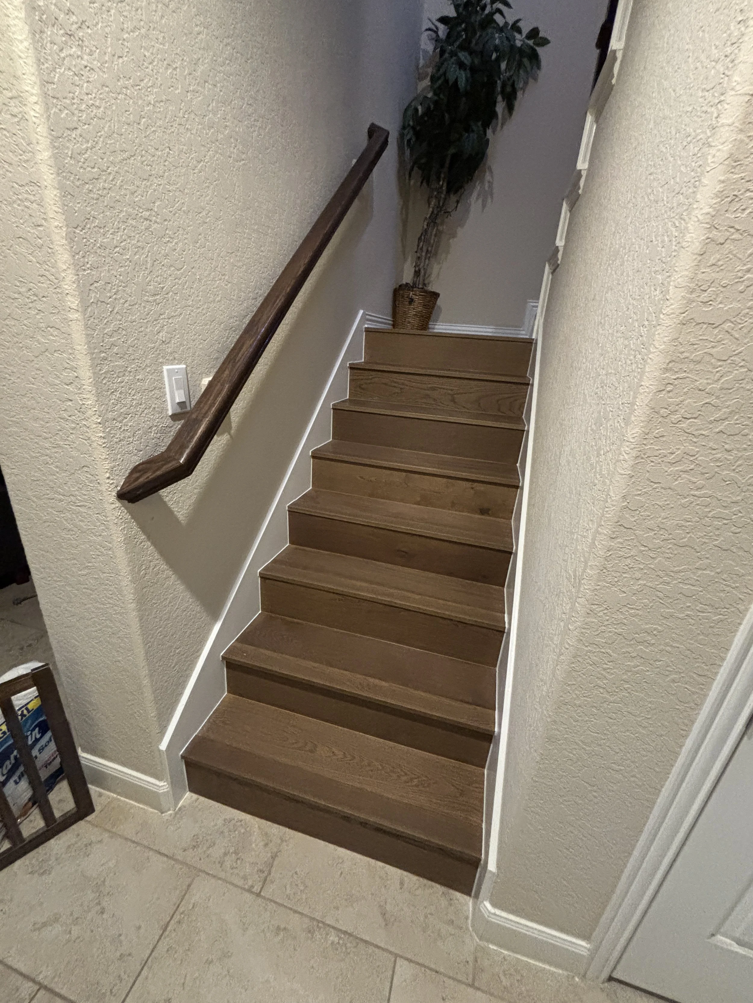 Indoor staircase with wooden steps, beige textured walls, a dark brown wooden handrail on the left, a potted plant at the top of the stairs, and a light switch on the wall.