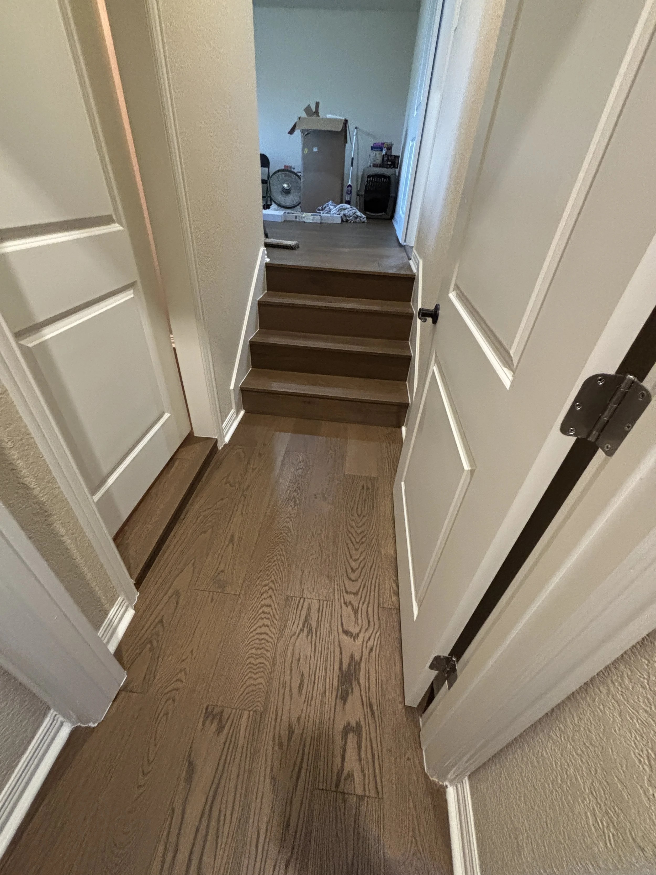 A view of a hallway with hardwood floors, leading to a room with cardboard boxes, a fan, and various household items.