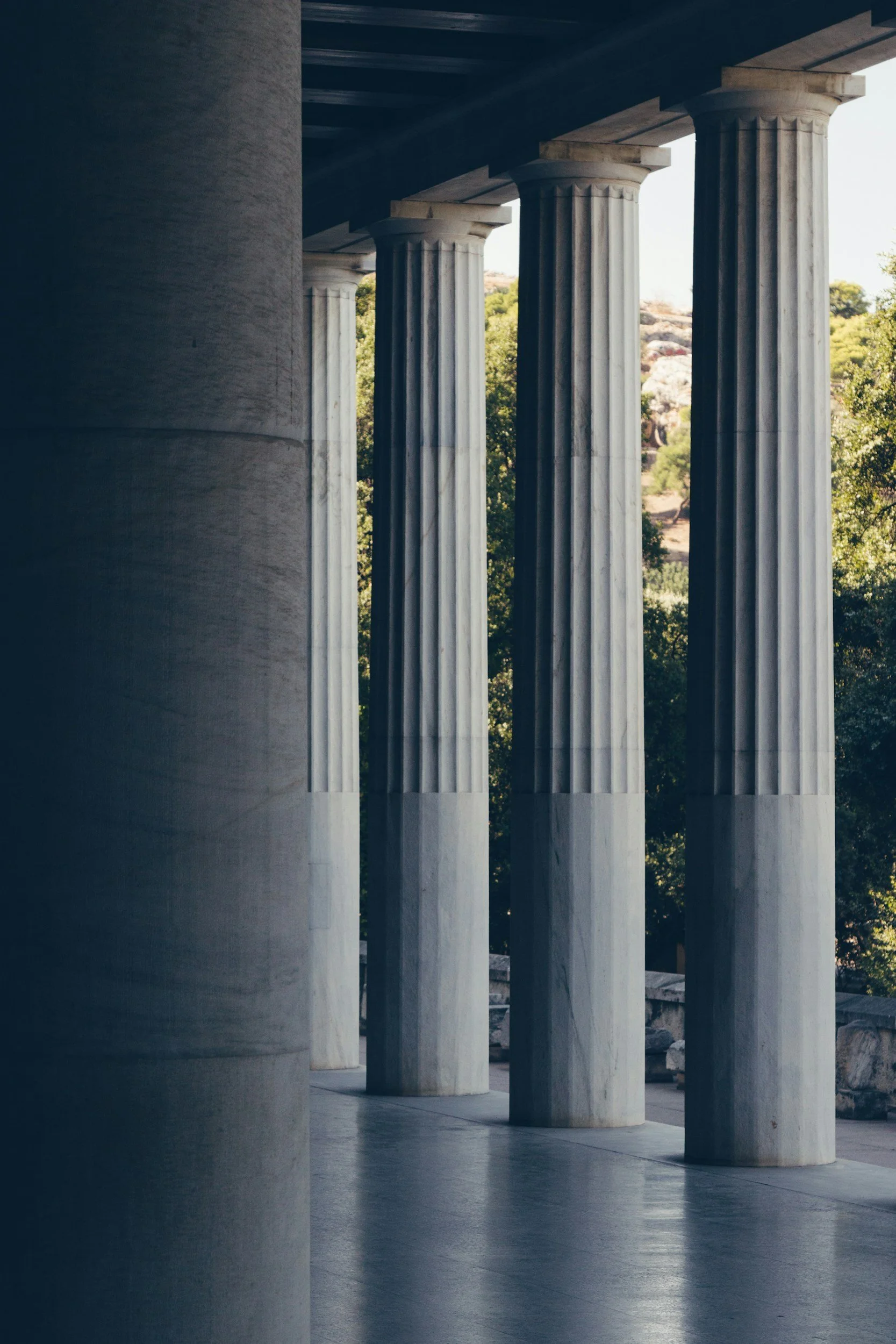 Ancient Greek-style marble columns in a row, supporting a structure, with trees and rocks visible in the background.