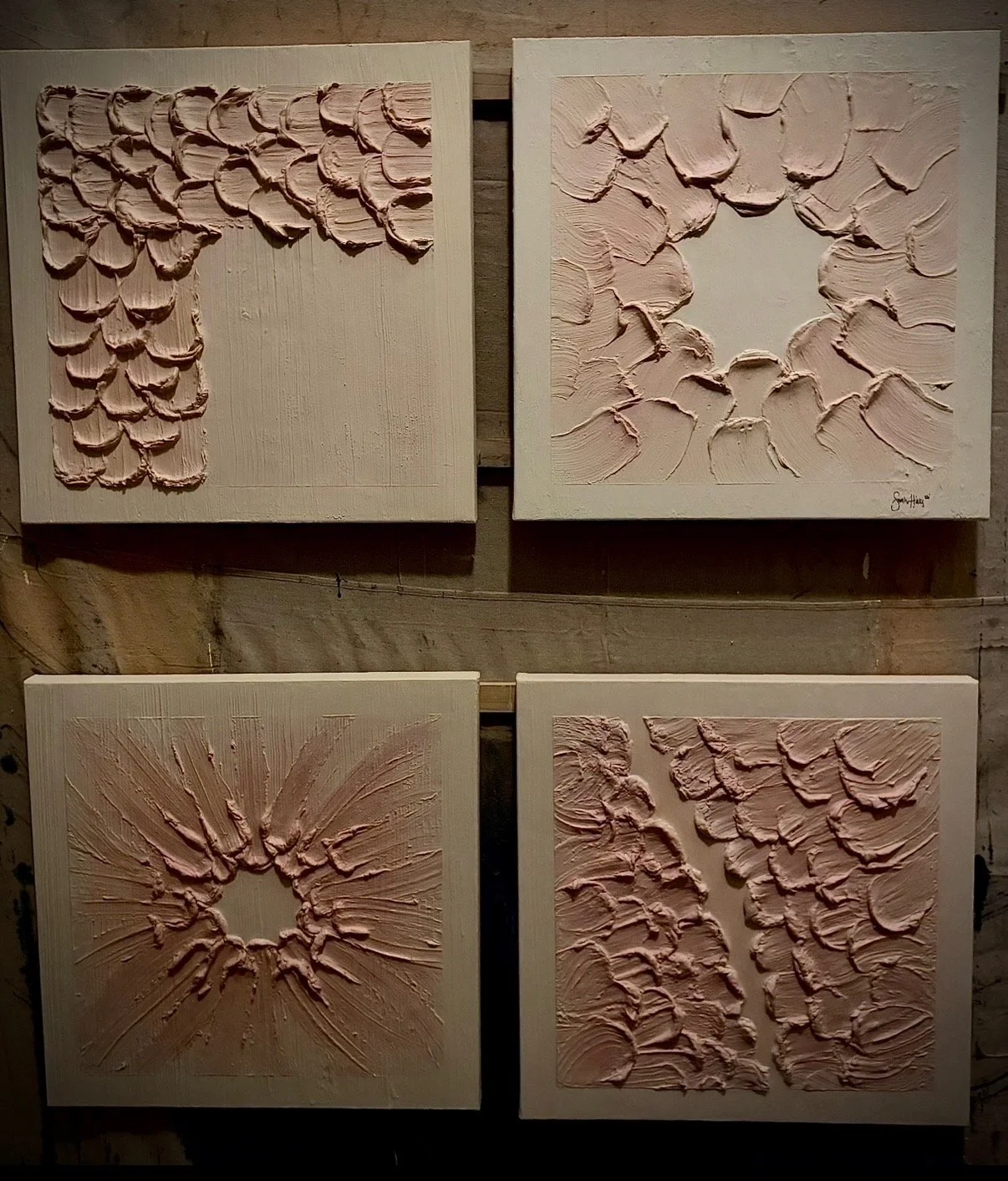 Four textured abstract paintings with pinkish paint, mounted on a wooden wall. Each feature bold, raised brushstrokes in various patterns, some circular and some linear.