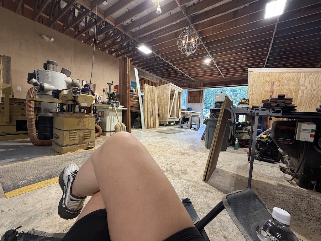 View from a person sitting with legs crossed in a woodworking workshop with various tools, wood pieces, and machinery, and a partially constructed wooden frame.