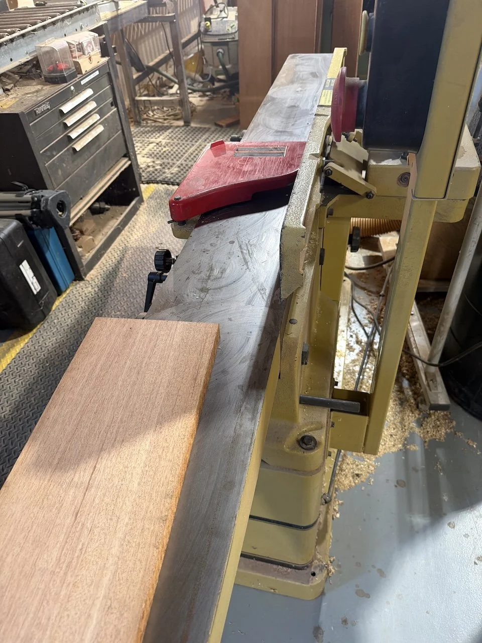 A woodworking workshop with a yellow benchtop thickness planer and a piece of wood on it, surrounded by woodworking tools and sawdust.