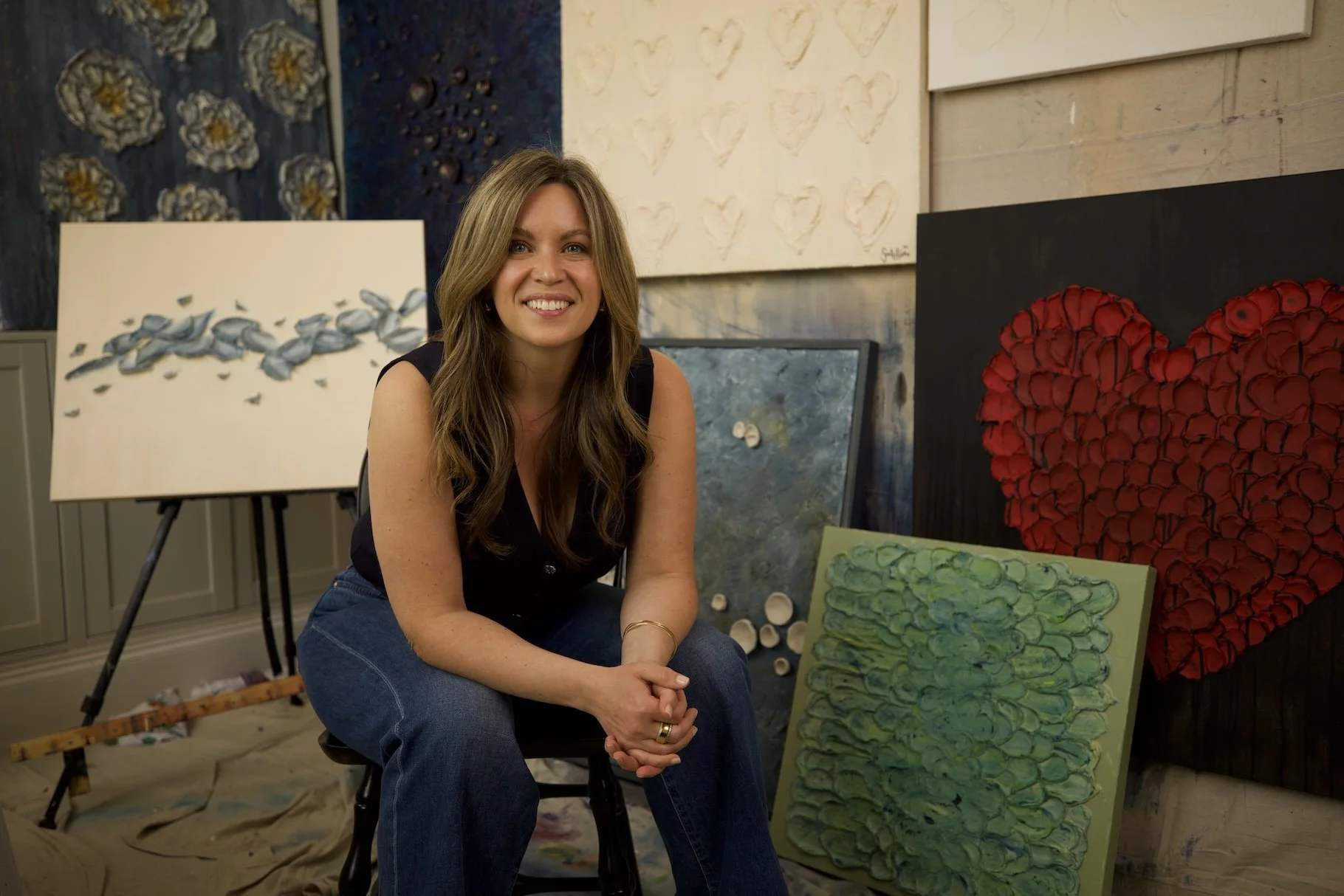 A woman with long wavy hair smiling in an art studio surrounded by abstract paintings, including a heart-shaped piece, a canvas with textured circles, and a painting with flying fish.